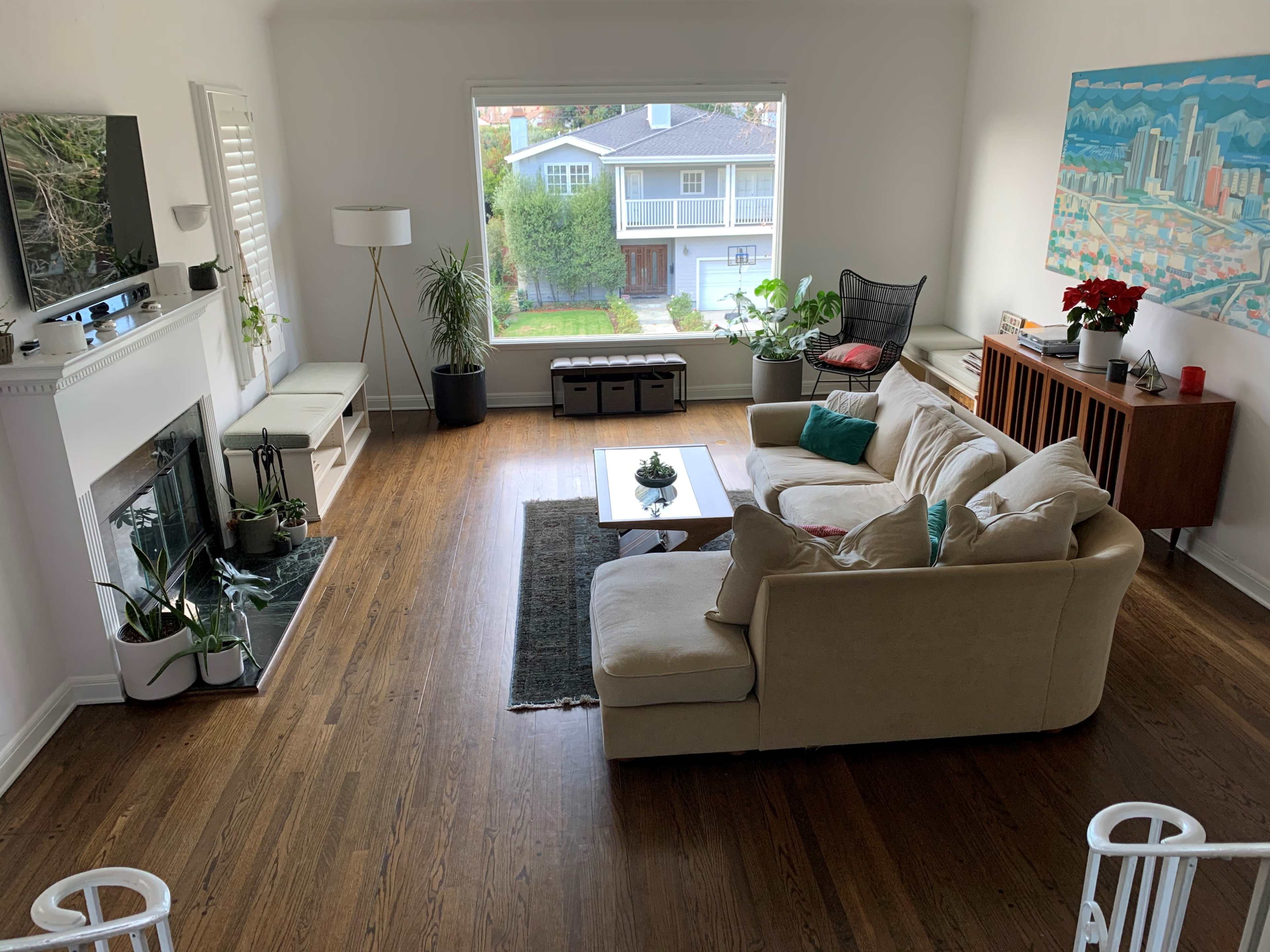 A spacious living room features a beige sofa, a coffee table, a flat-screen TV mounted above a fireplace, and a large window overlooking a garden area.