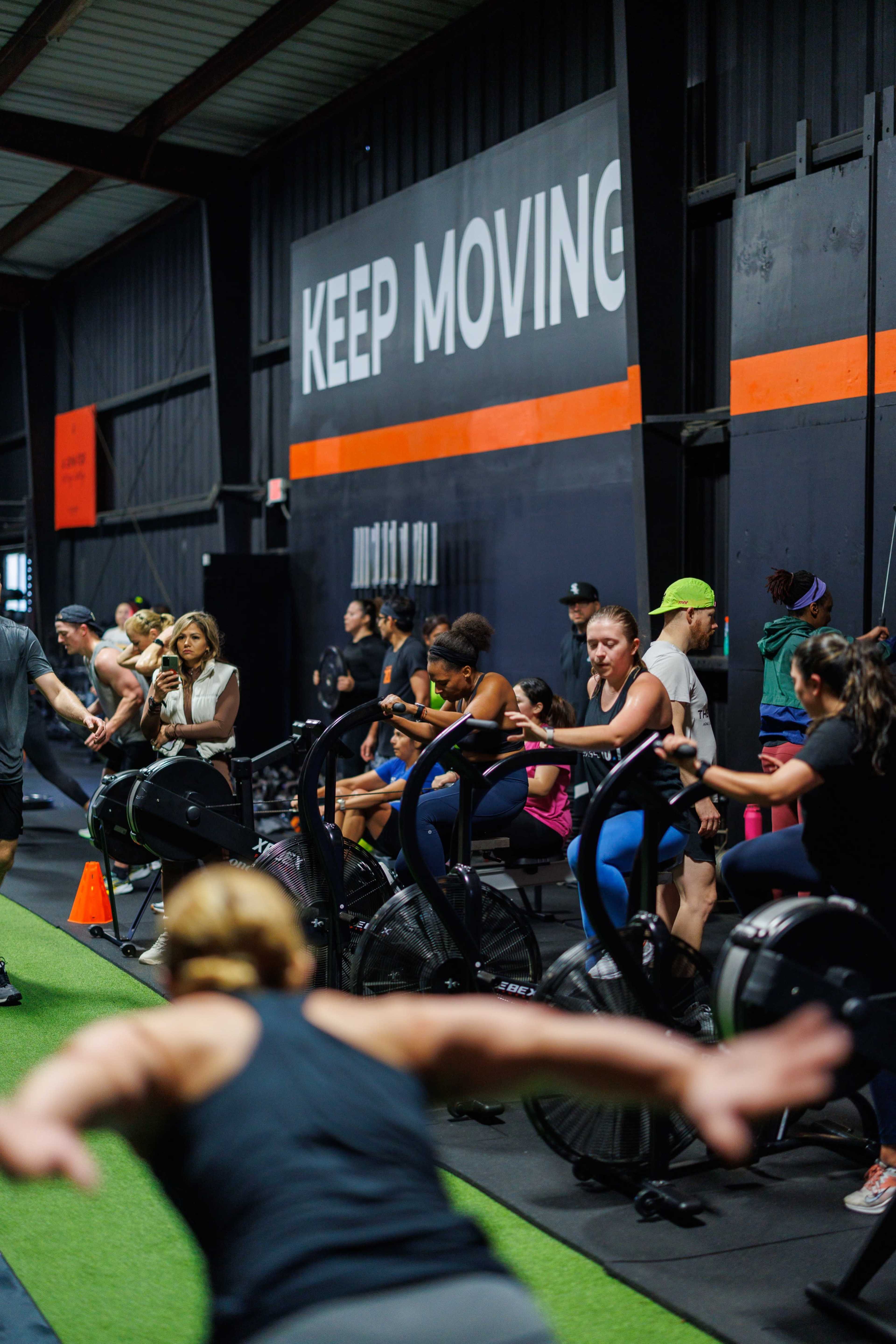 A group of people is engaged in a workout session at a fitness facility, using exercise bikes and rowing machines under a "KEEP MOVING" wall sign.