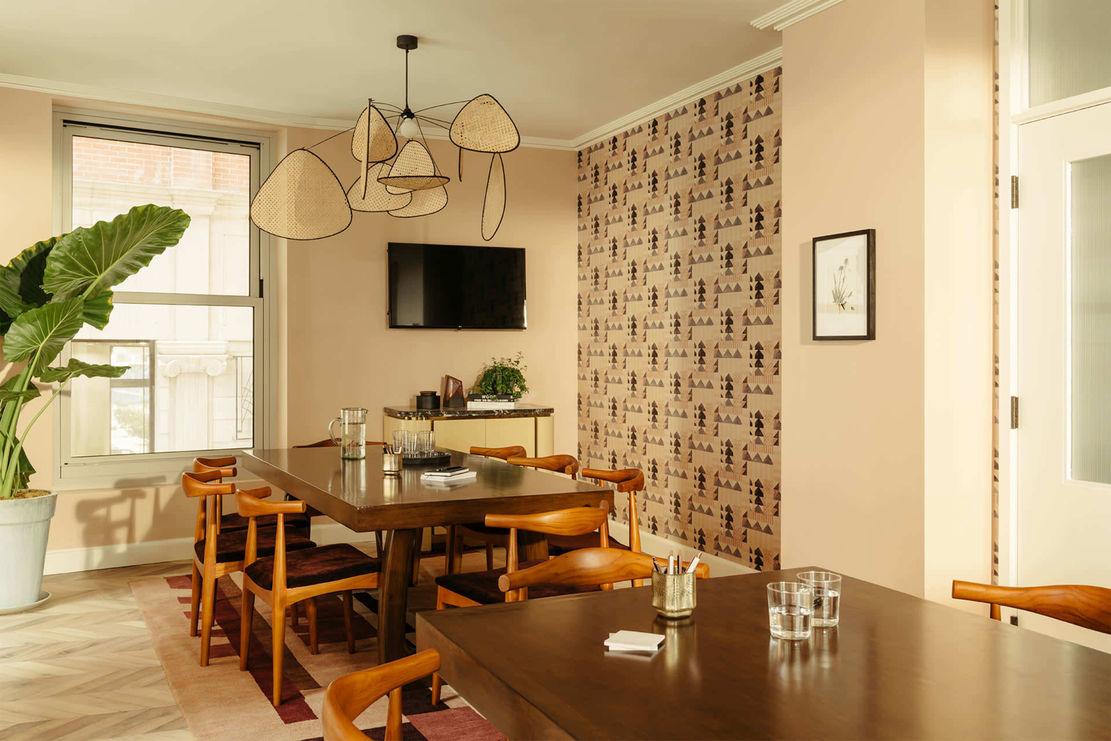 The image shows a modern dining area with wooden tables and chairs, a television mounted on the wall, and a large potted plant near the window.