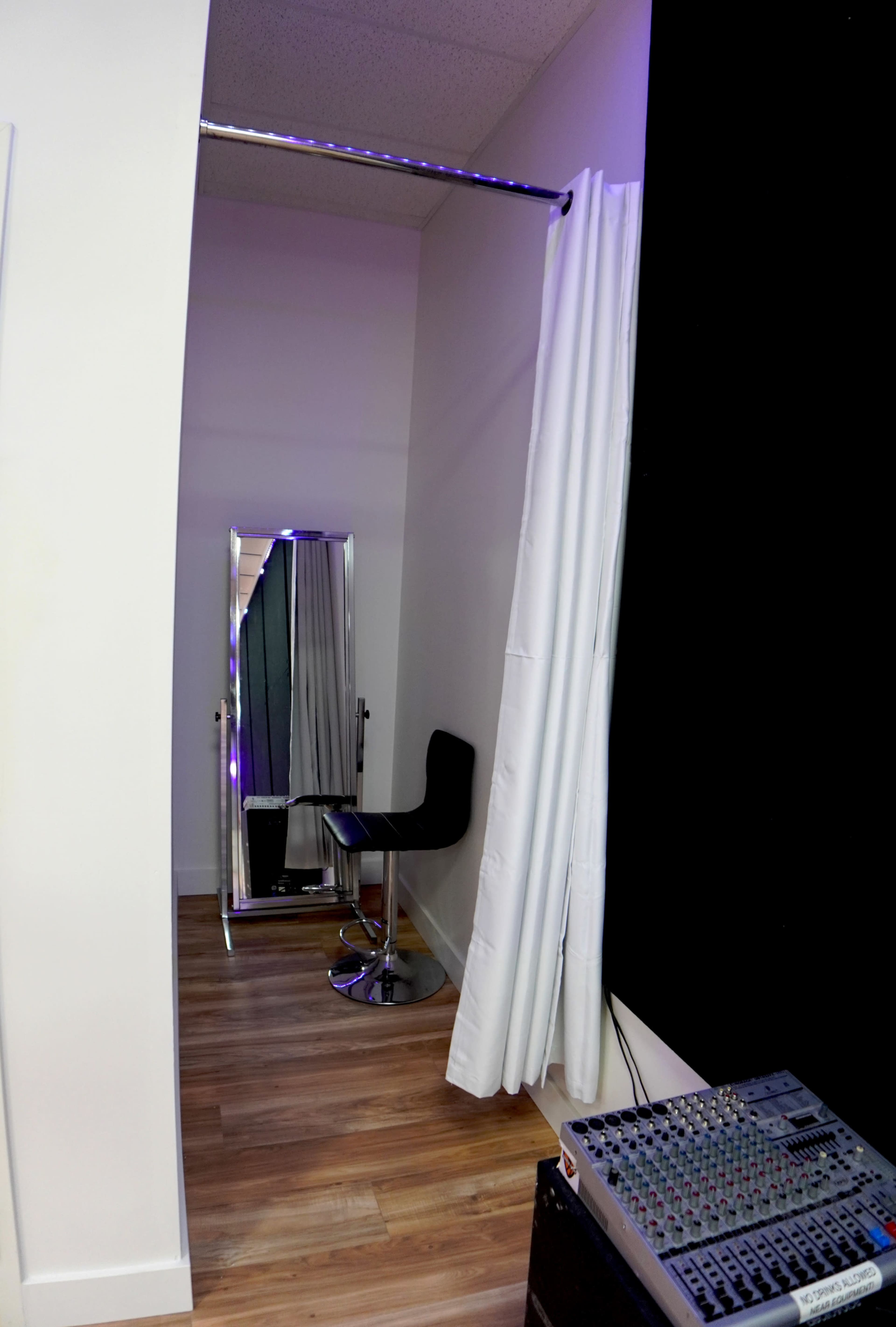 A small room with a mirrored wall, a black stool, white curtains, and an audio mixing console in the corner.