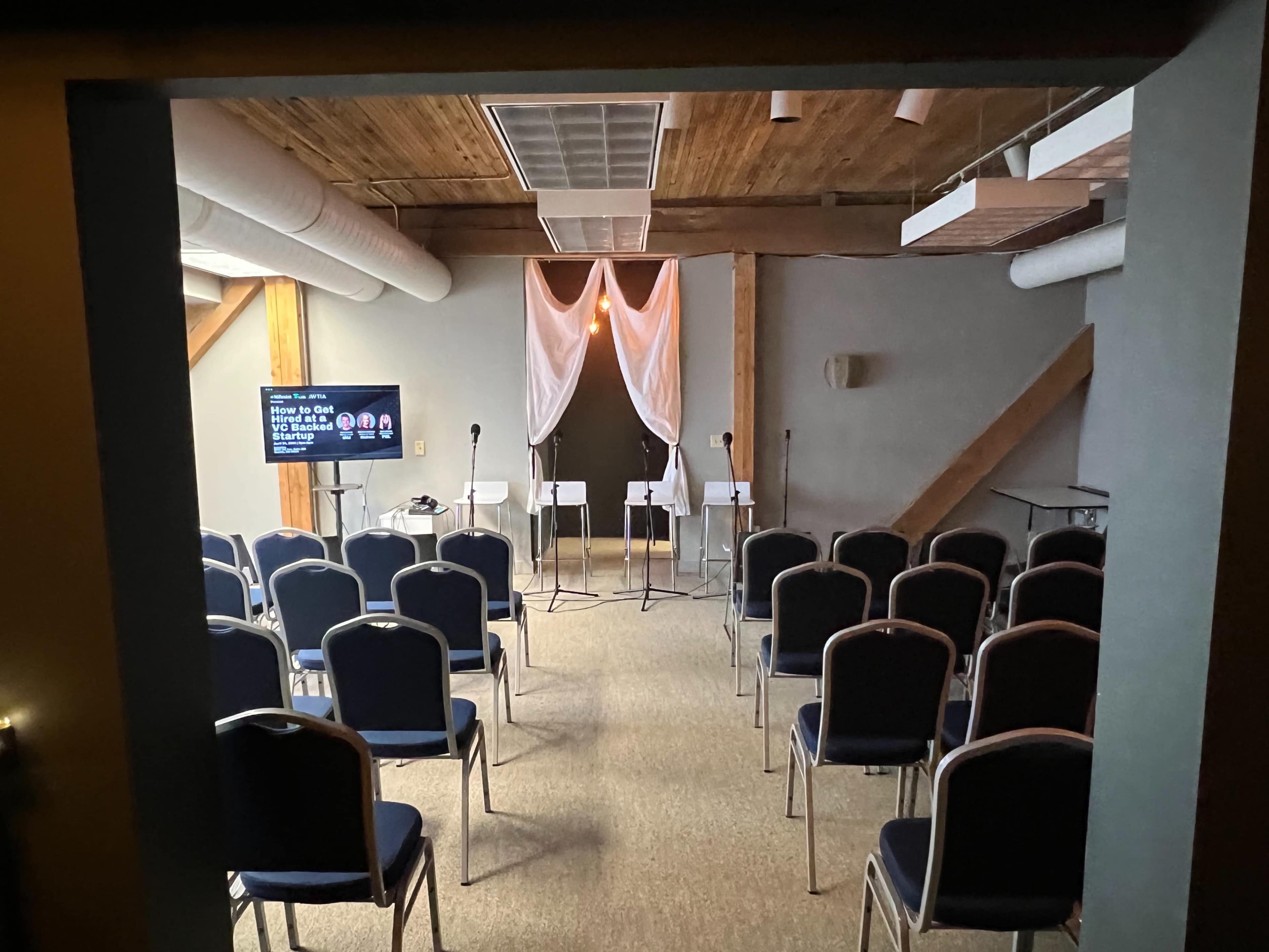 A small indoor event space with rows of chairs facing a stage area that has three microphones and a screen displaying text.