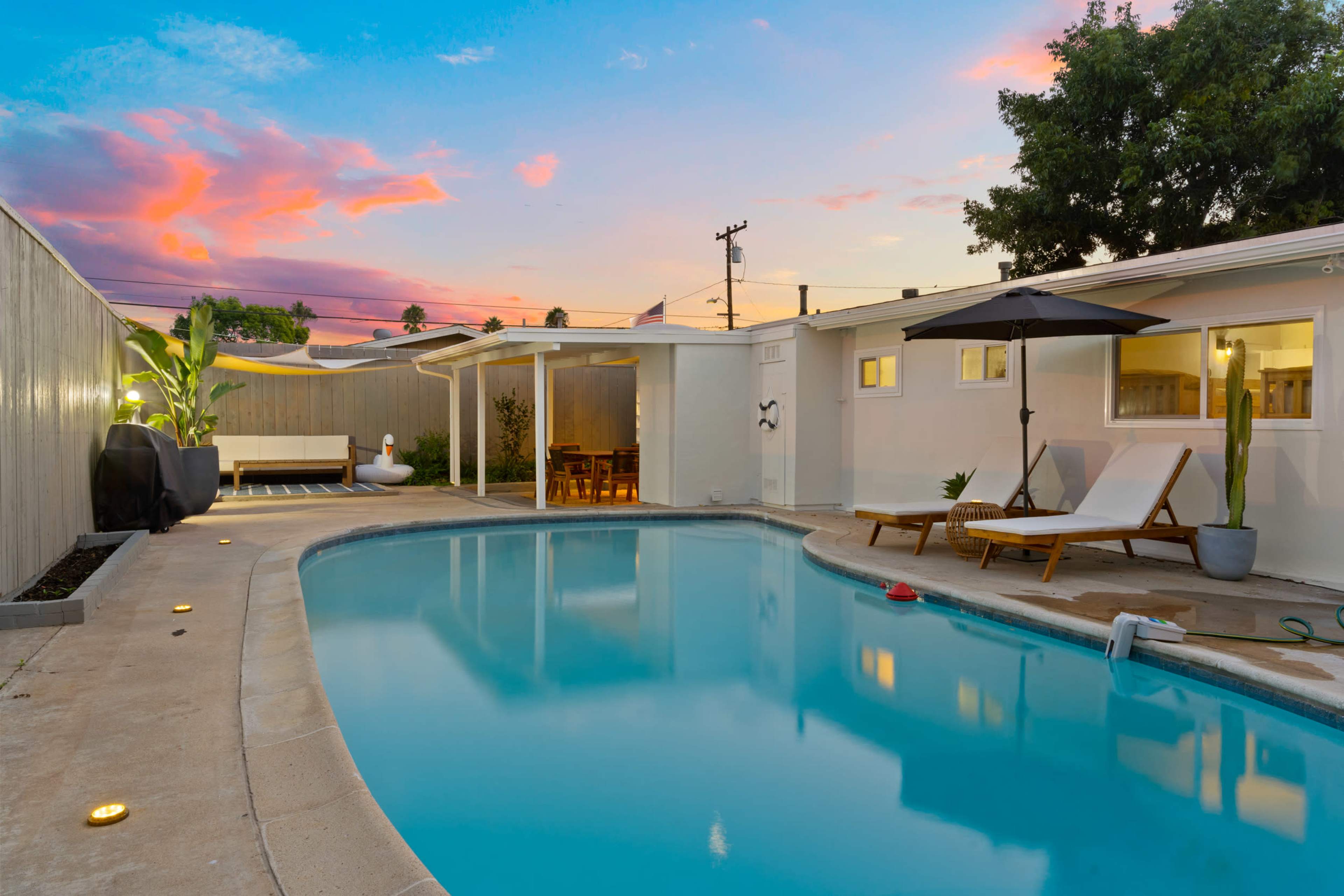 A swimming pool area surrounded by lounge chairs and plants, with a pink and blue sky at sunset.