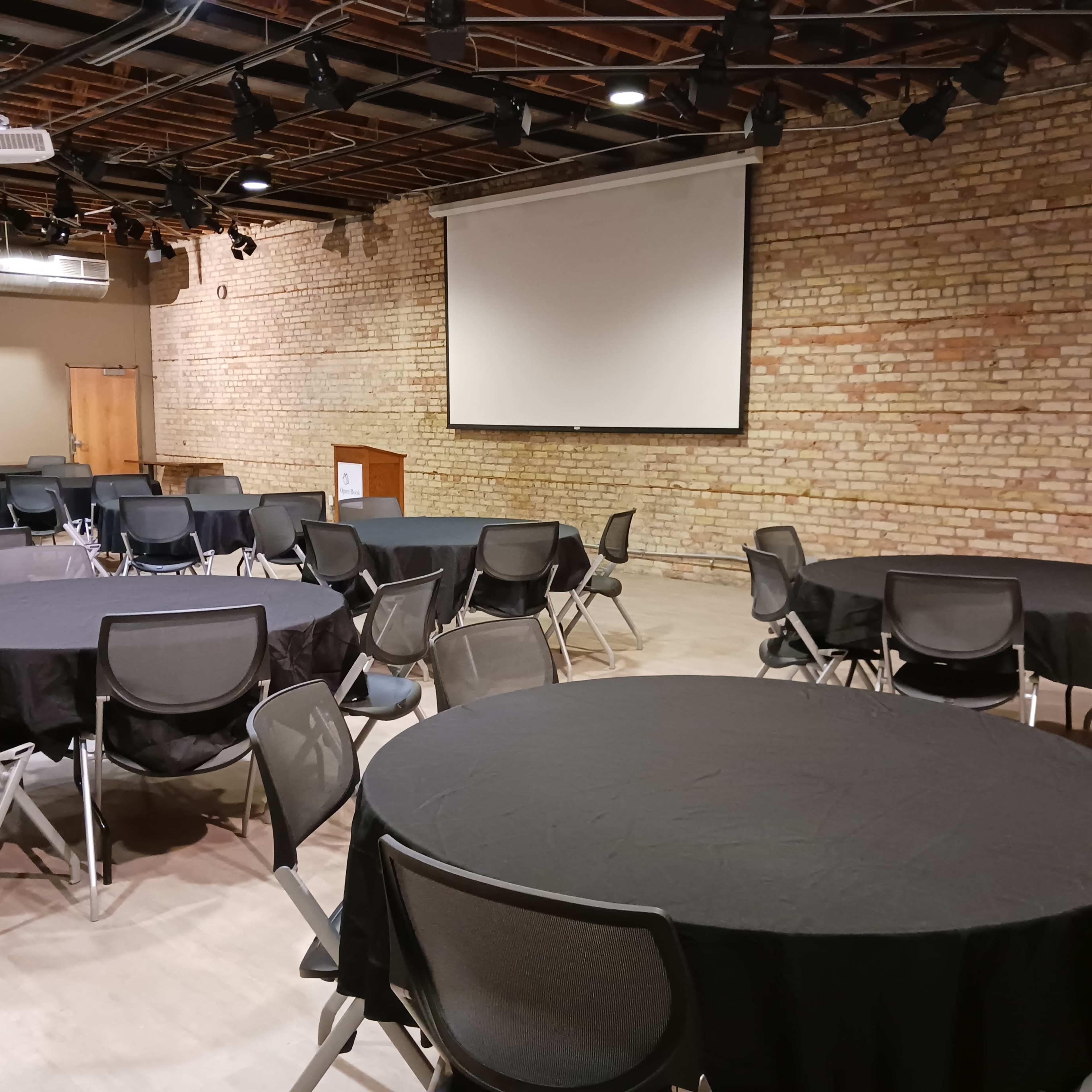A spacious event room features several round tables covered with black tablecloths, with a large blank projection screen on the wall.