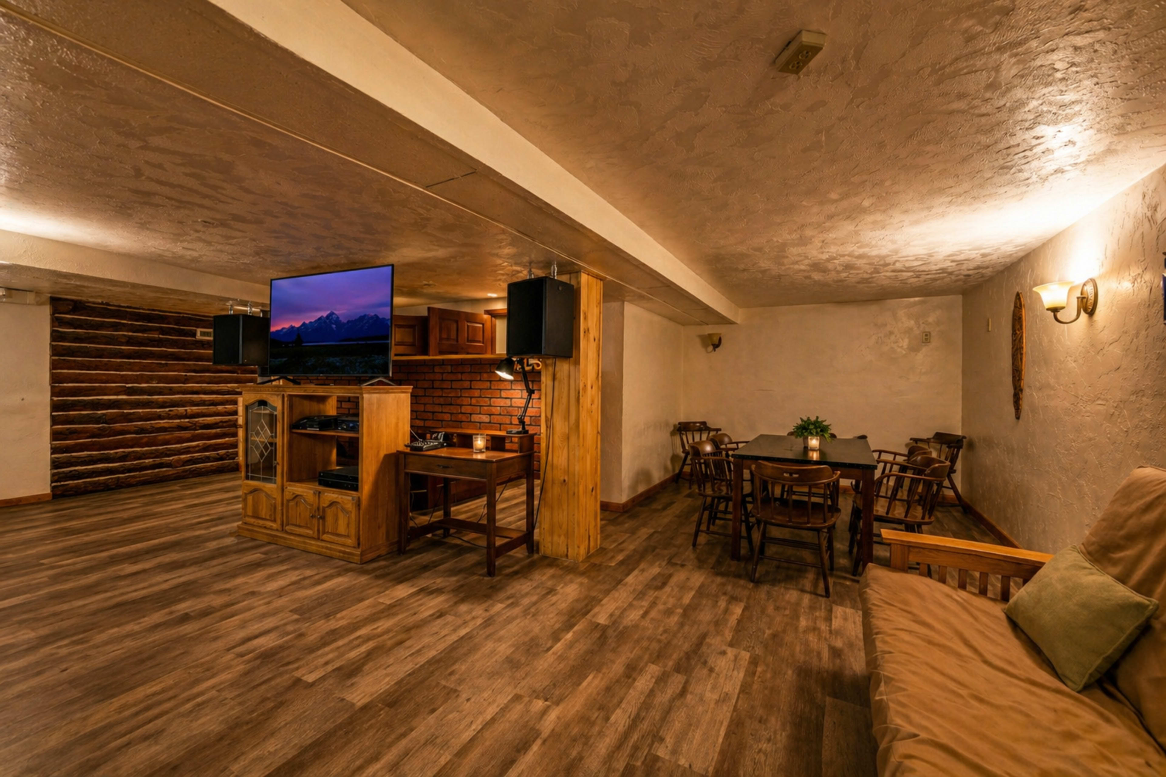 The image shows a spacious basement room featuring a wooden dining table, a couch, a television mounted on the wall, and log accents throughout the space.