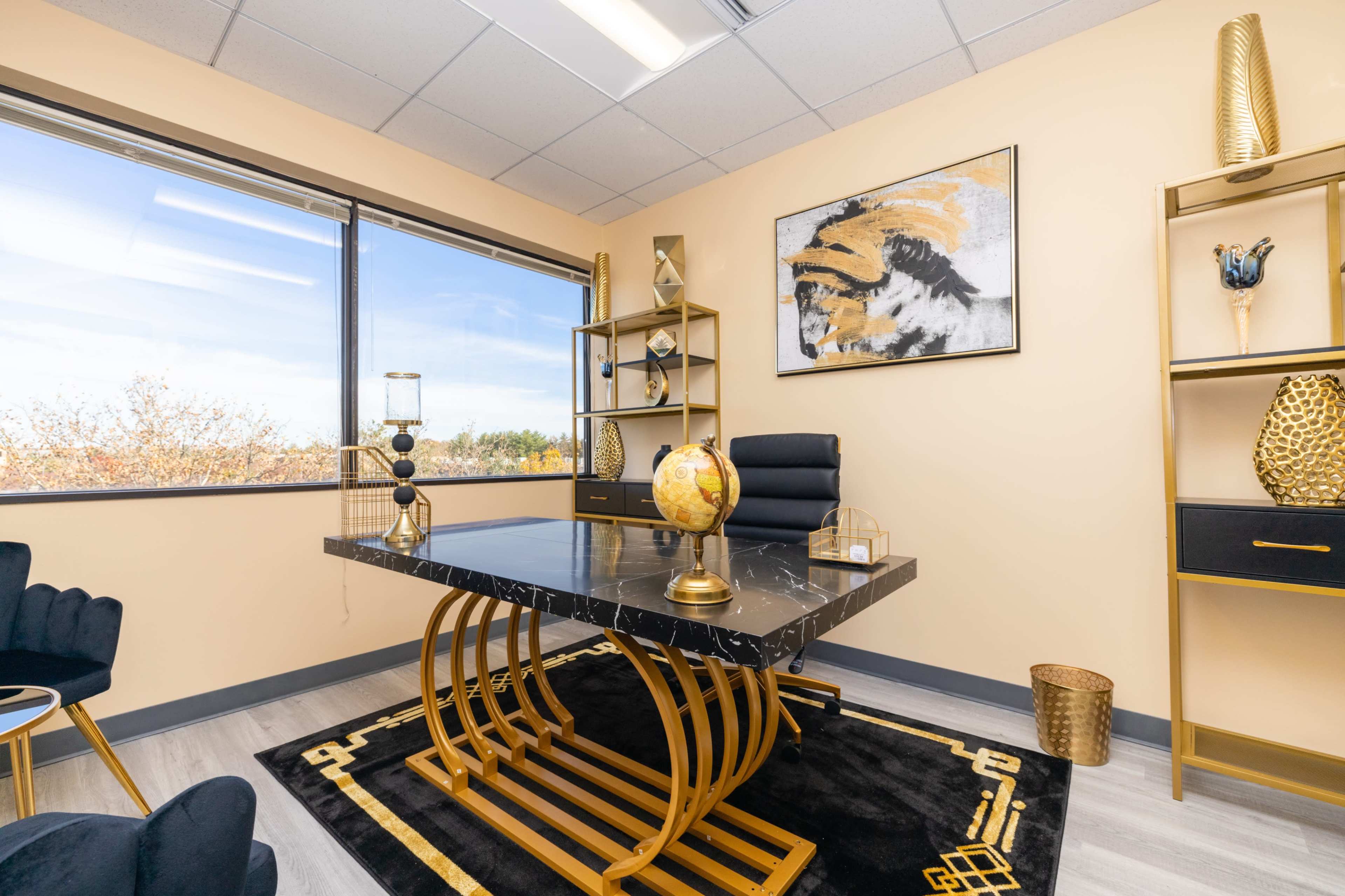 A modern office features a black marble desk with a gold base, surrounded by black chairs and decorative shelves against a window with a view of trees outside.