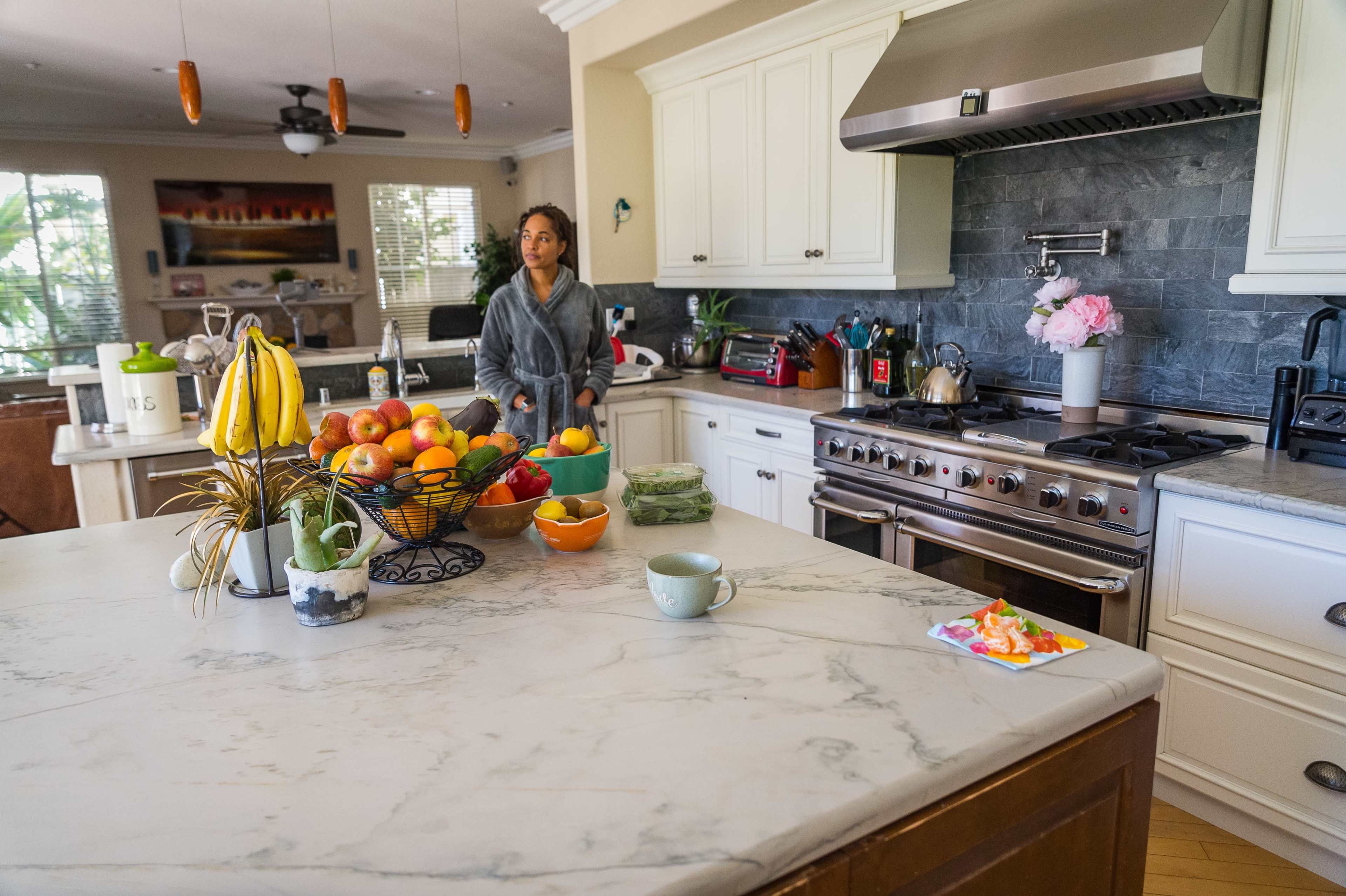 A person stands in a kitchen with a marble countertop, surrounded by bowls of fresh fruit and a bowl of vegetables, near a stove and various kitchen appliances.
