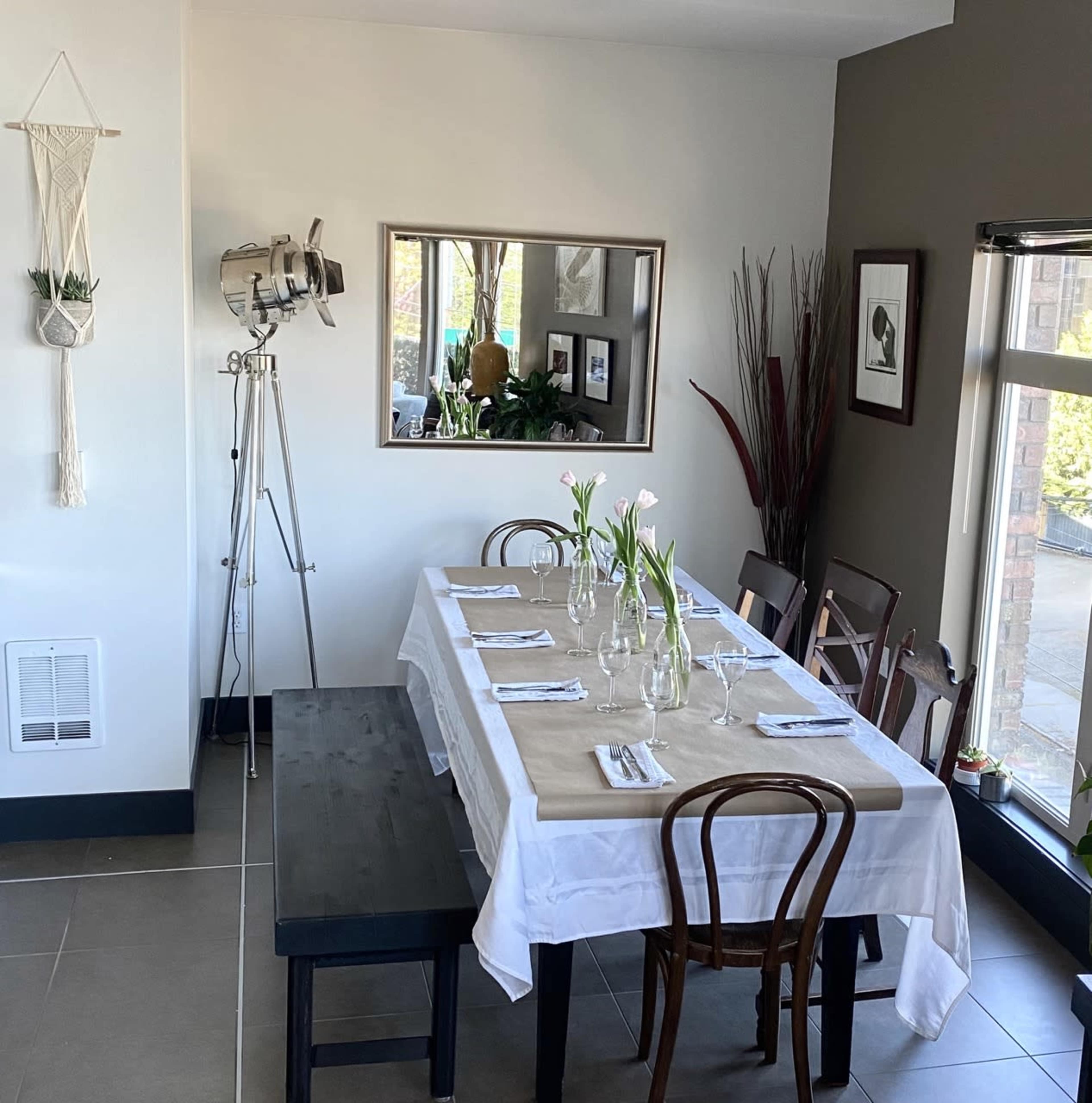 A dining area features a long table set with glassware and flowers, surrounded by chairs and a decorative mirror on the wall.