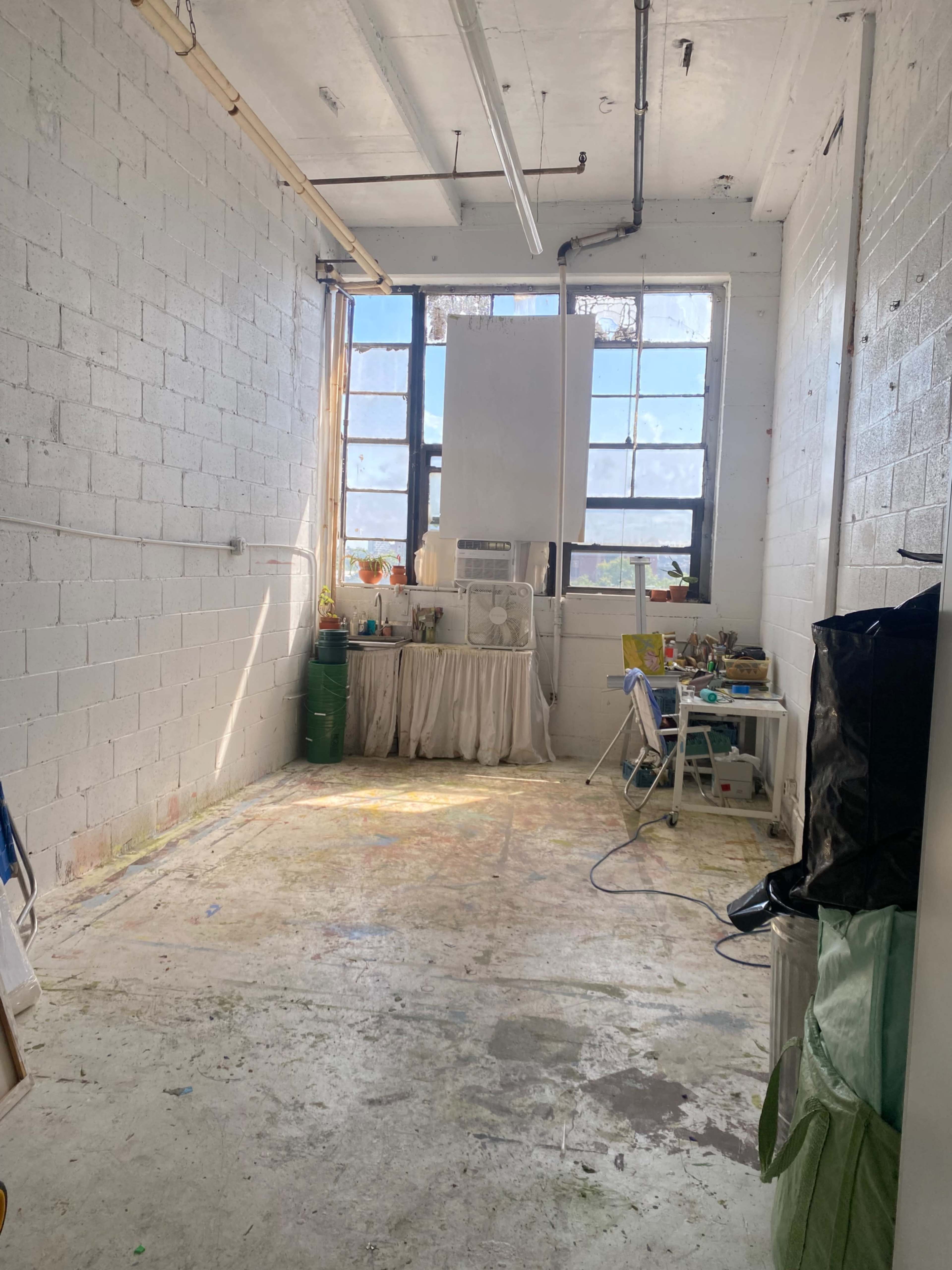 The image shows a spacious room with bare walls, large windows allowing natural light, and a corner setup with a fan, table, and other items.