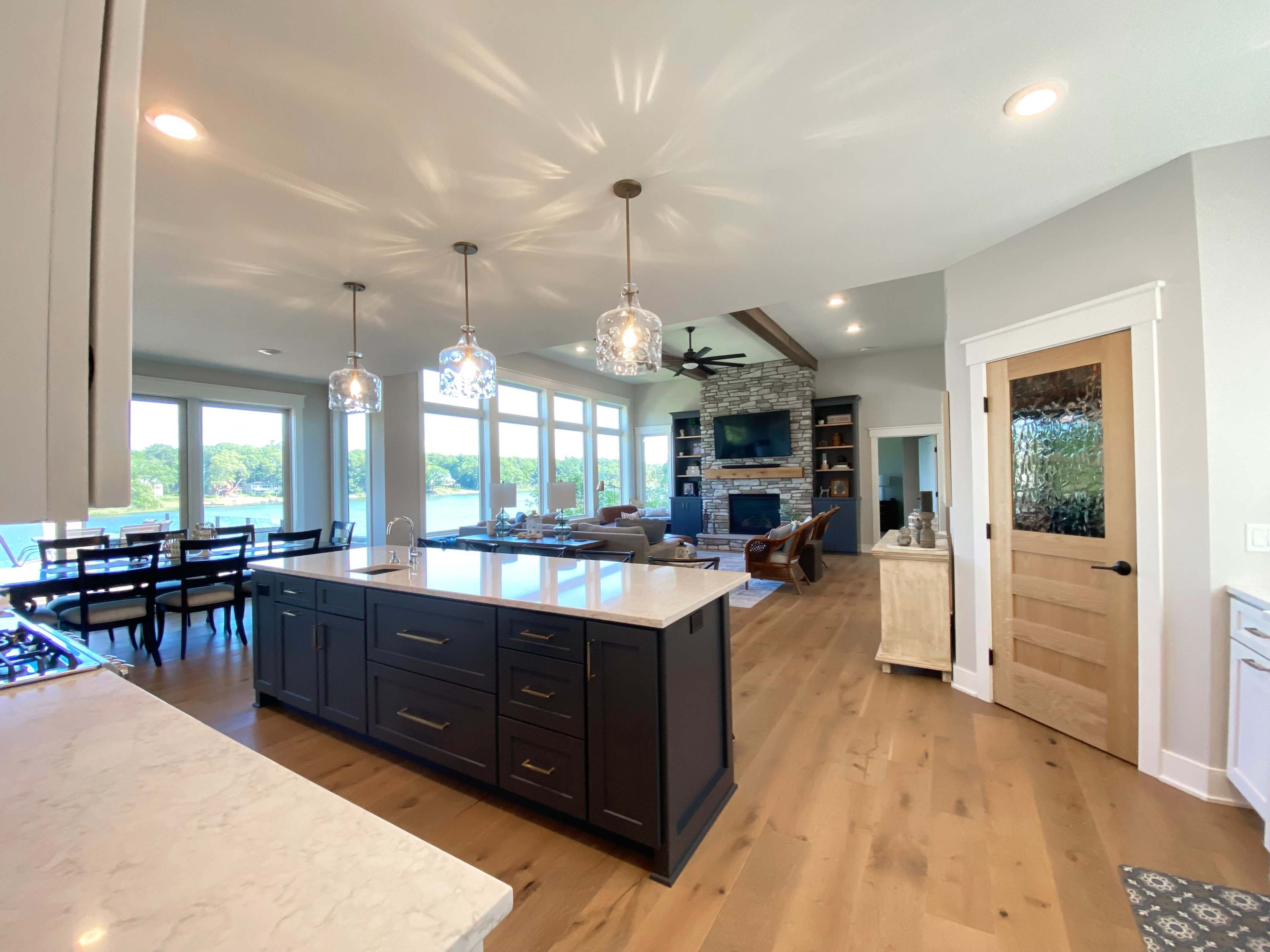 A modern kitchen features a large island with dark cabinetry and pendant lights, leading to an open living area with large windows overlooking a scenic view.