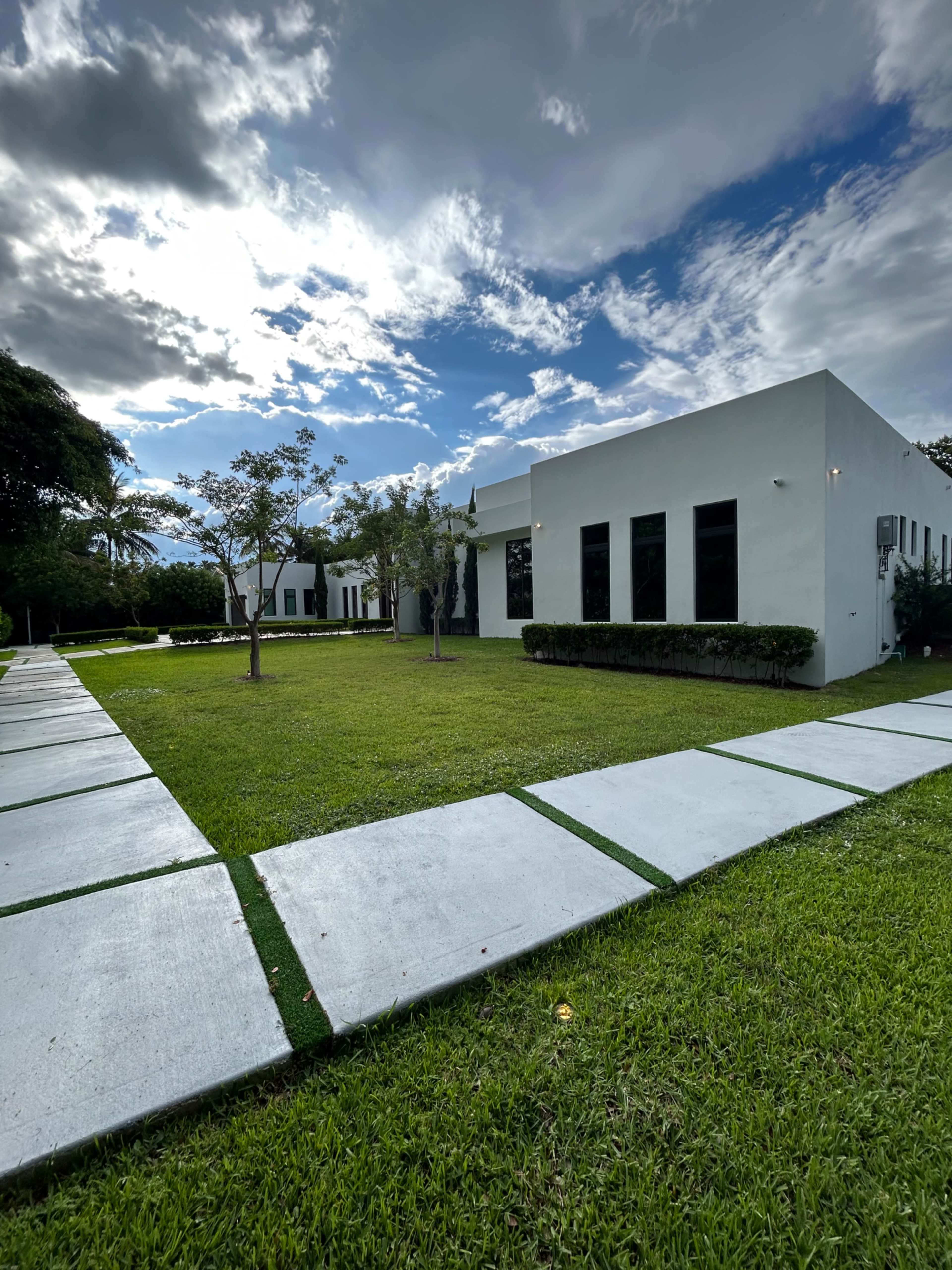 A modern, minimalist white building surrounded by manicured grass and a concrete pathway under a partly cloudy sky.