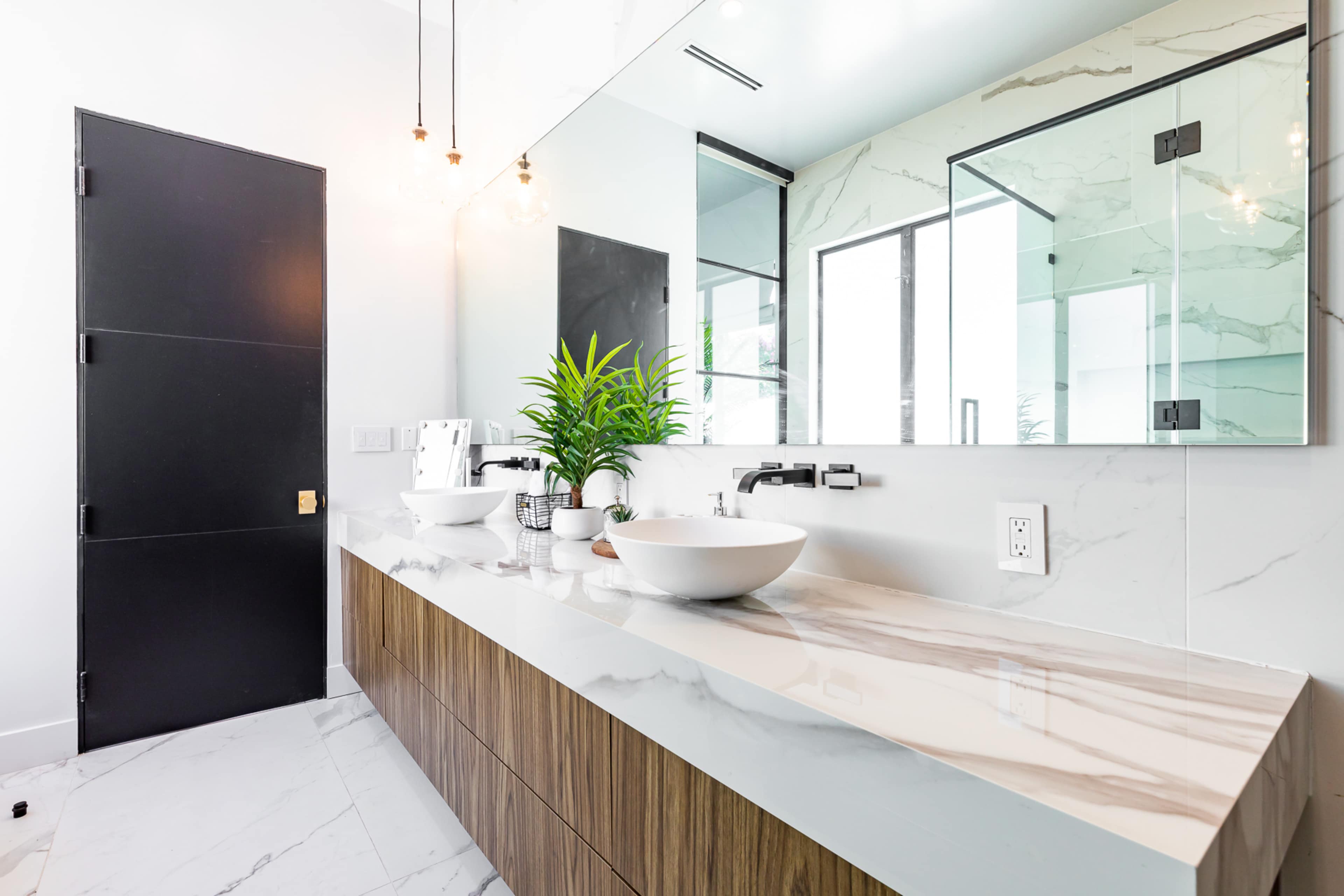 A modern bathroom with a wooden vanity, a large mirror, and a countertop featuring a round basin, surrounded by neatly arranged decor and plants.
