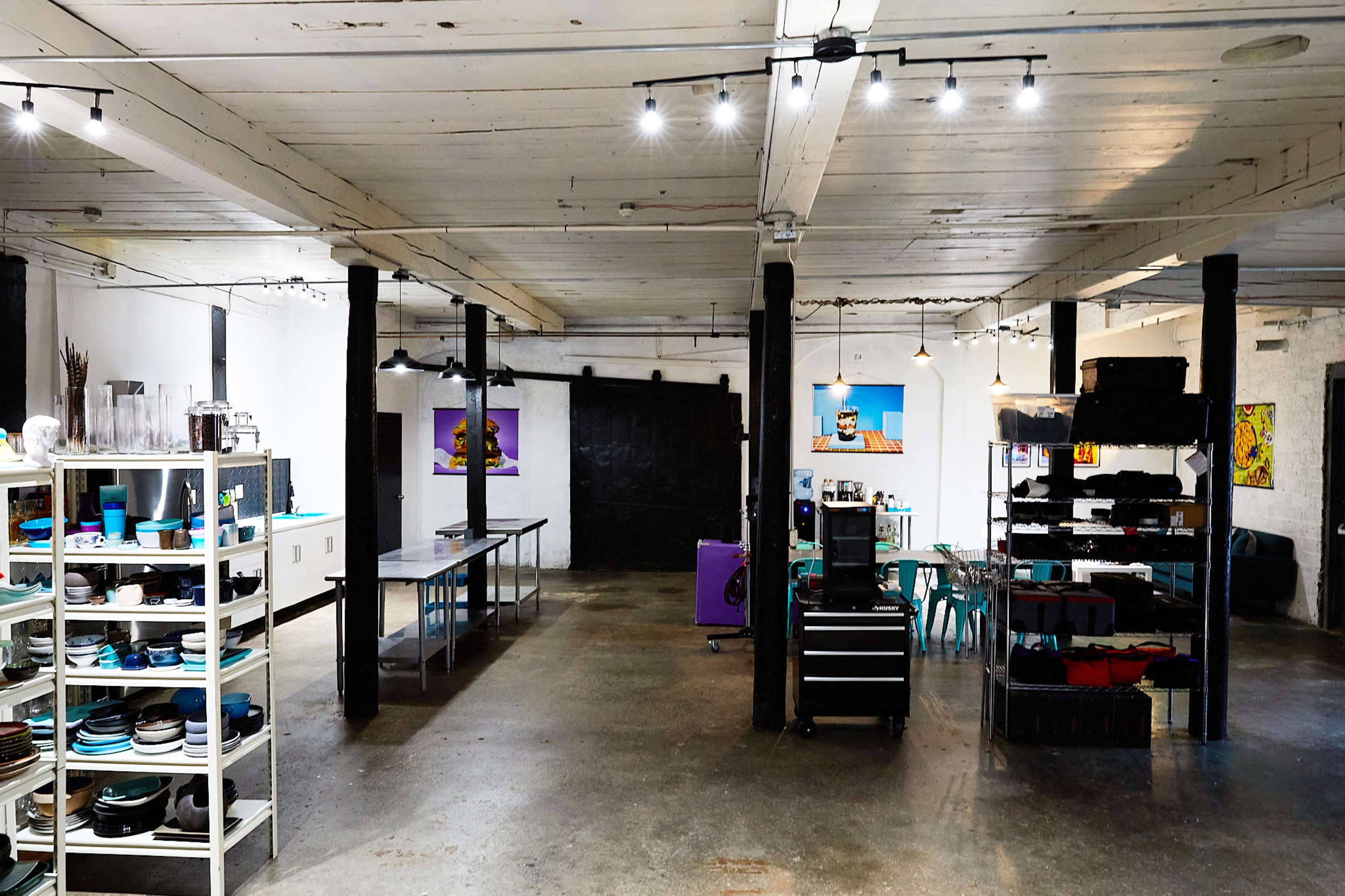 A spacious, industrial-style room features tables and shelves filled with various kitchen items, while art hangs on the walls and concrete flooring provides a minimalist backdrop.