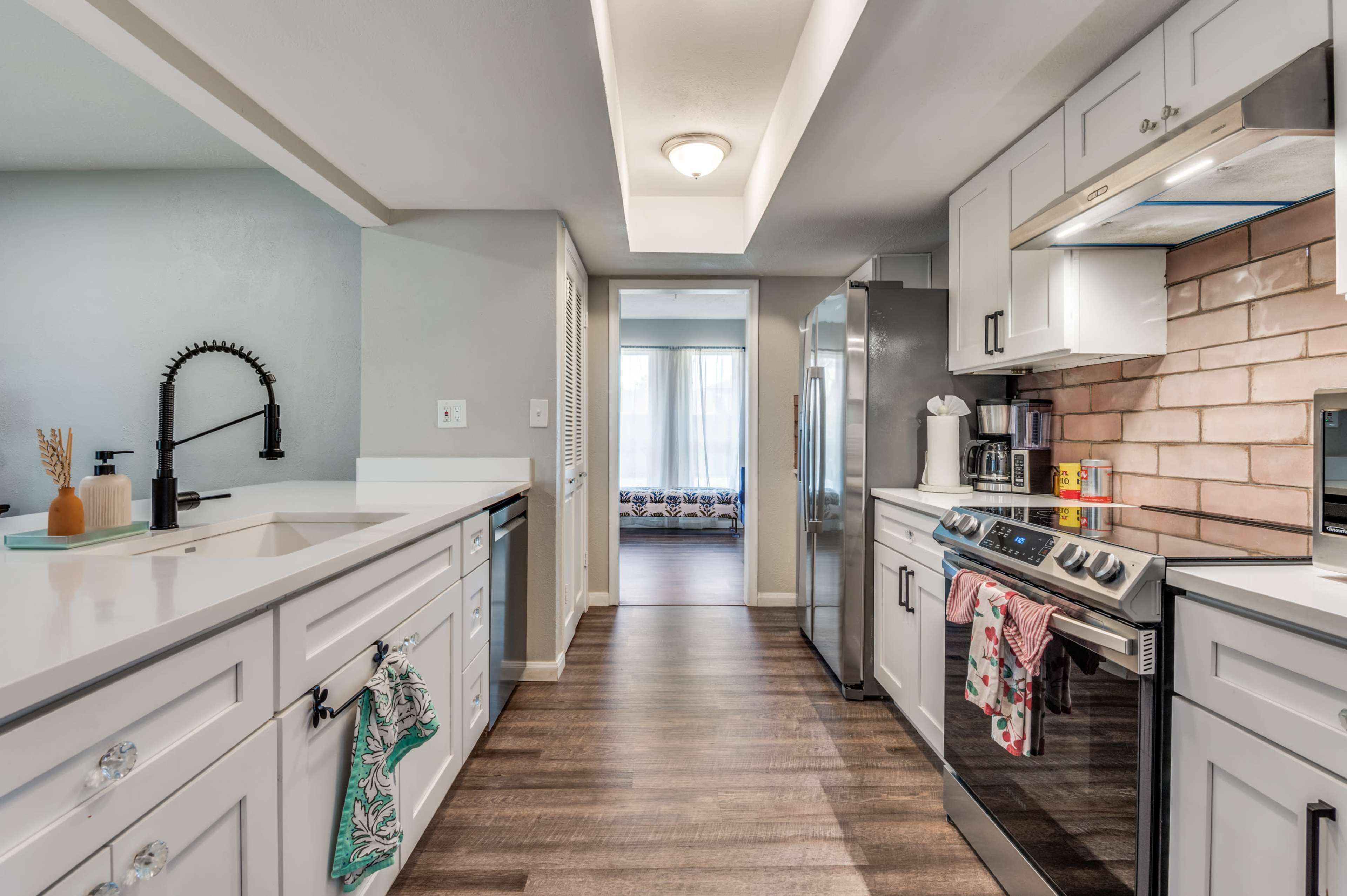 The image shows a modern kitchen with white cabinets, a stainless steel refrigerator, and a stove with a hood, leading to a hallway and a bright living area in the background.