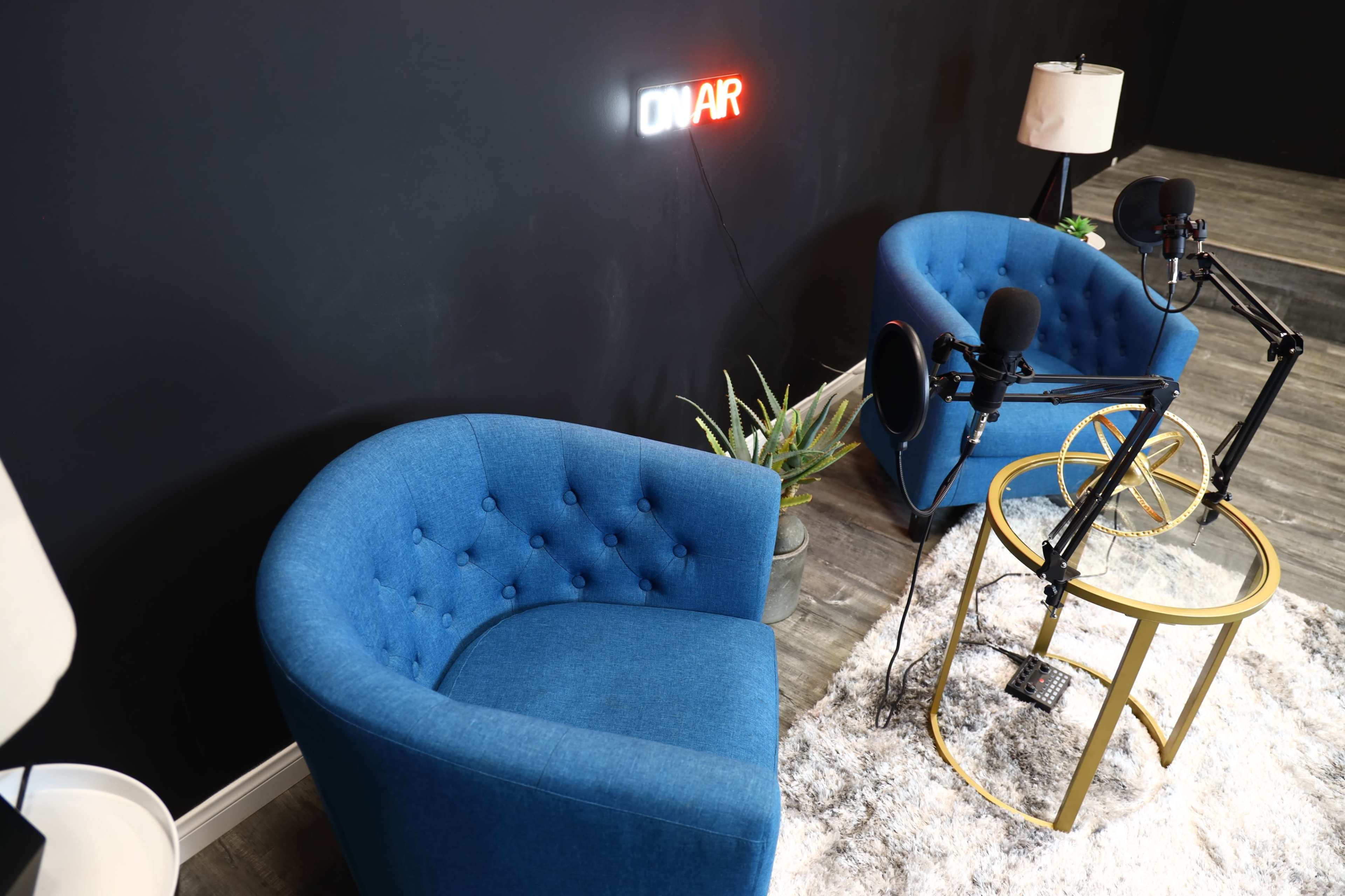 The image shows a small studio setup with two blue armchairs, a round table, microphones, and a neon "ON AIR" sign on a dark wall.