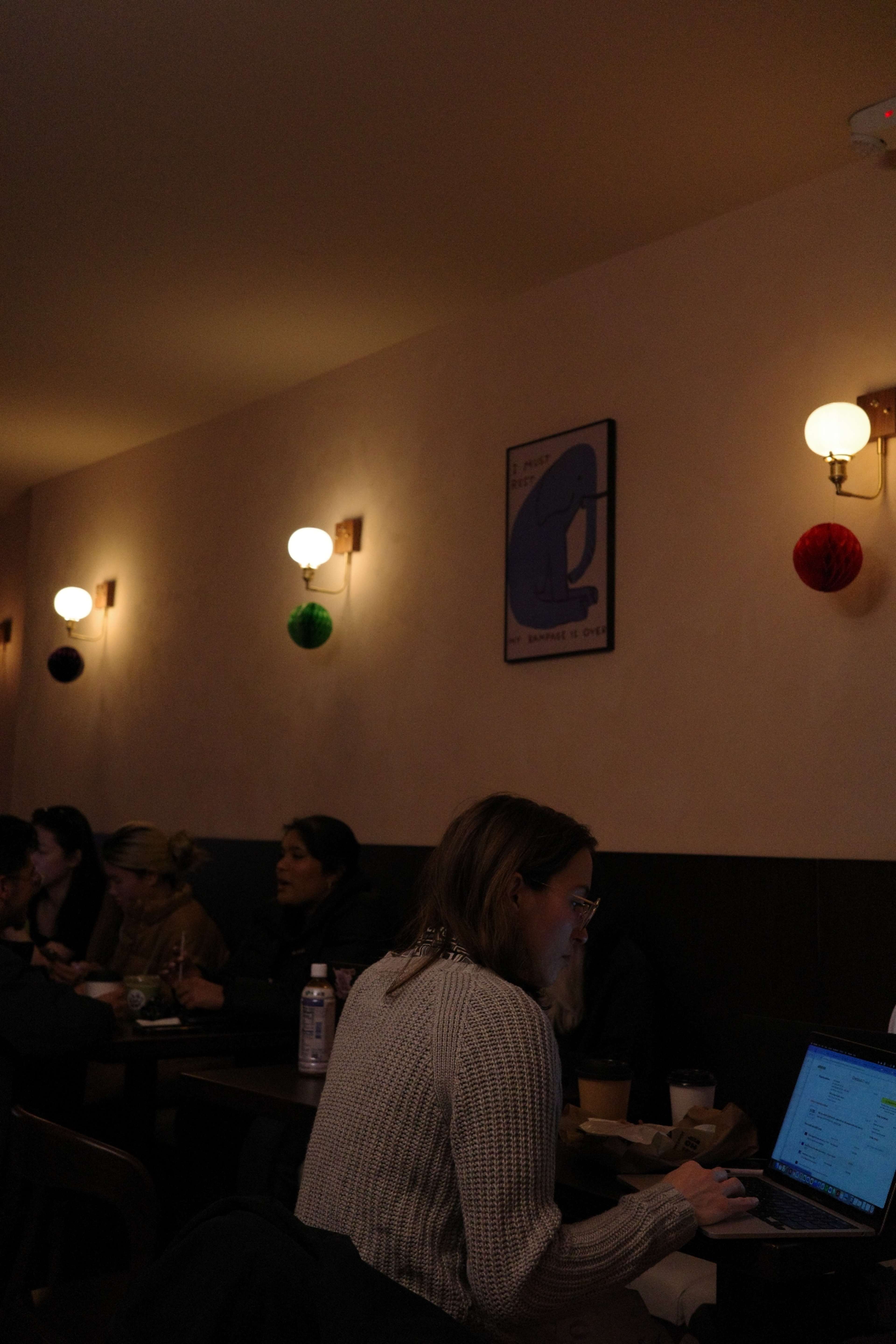 A woman works on a laptop at a café with several other patrons seated around her.