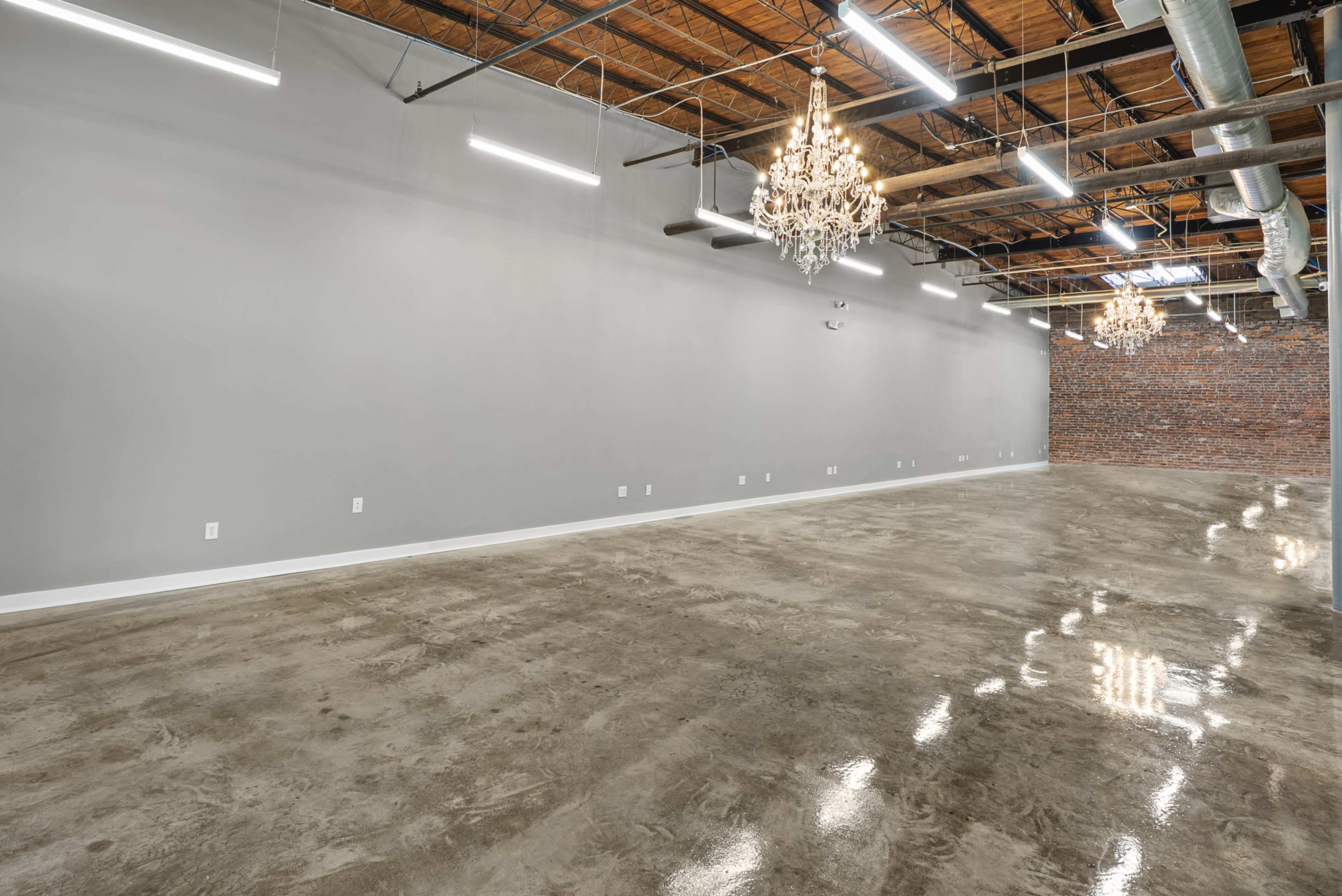 The image shows an empty industrial-style interior with polished concrete floors, exposed brick walls, and ornate chandeliers hanging from the ceiling.