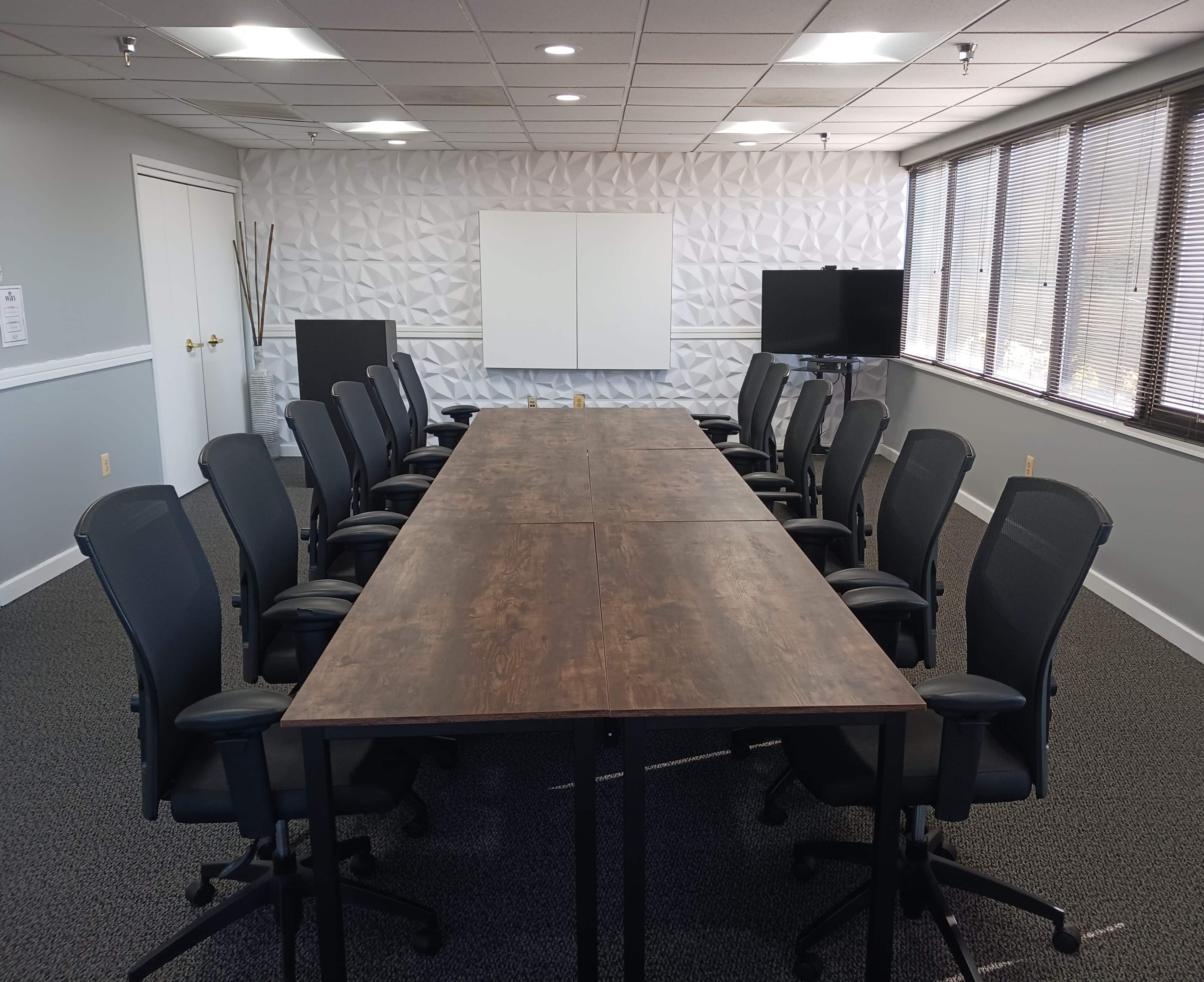 A conference room features a long wooden table with ten ergonomic chairs on each side, complemented by a whiteboard and a monitor at one end.