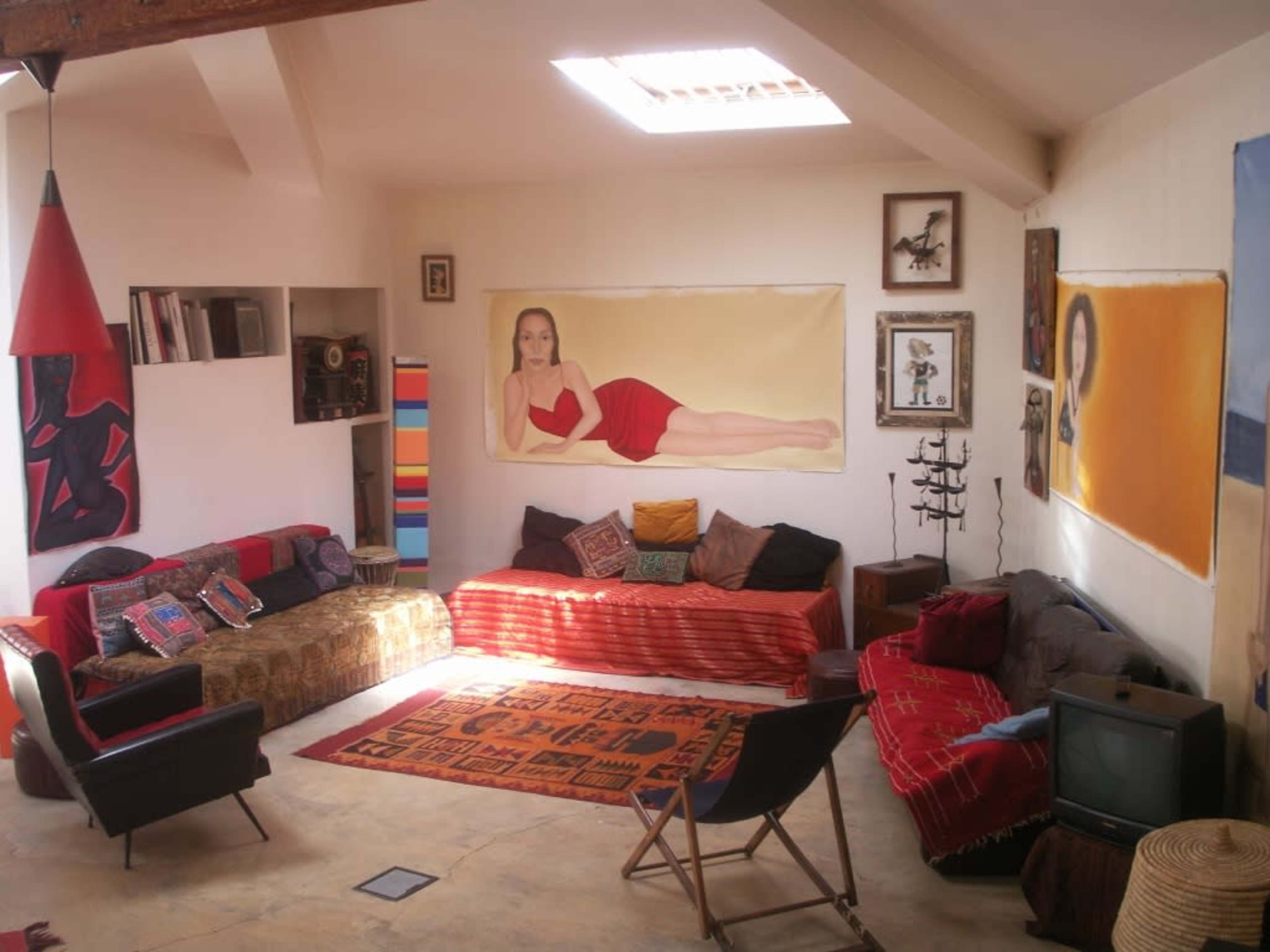 A sunlit living room featuring colorful couches, various wall art, and a mix of decorative pillows on the floor.