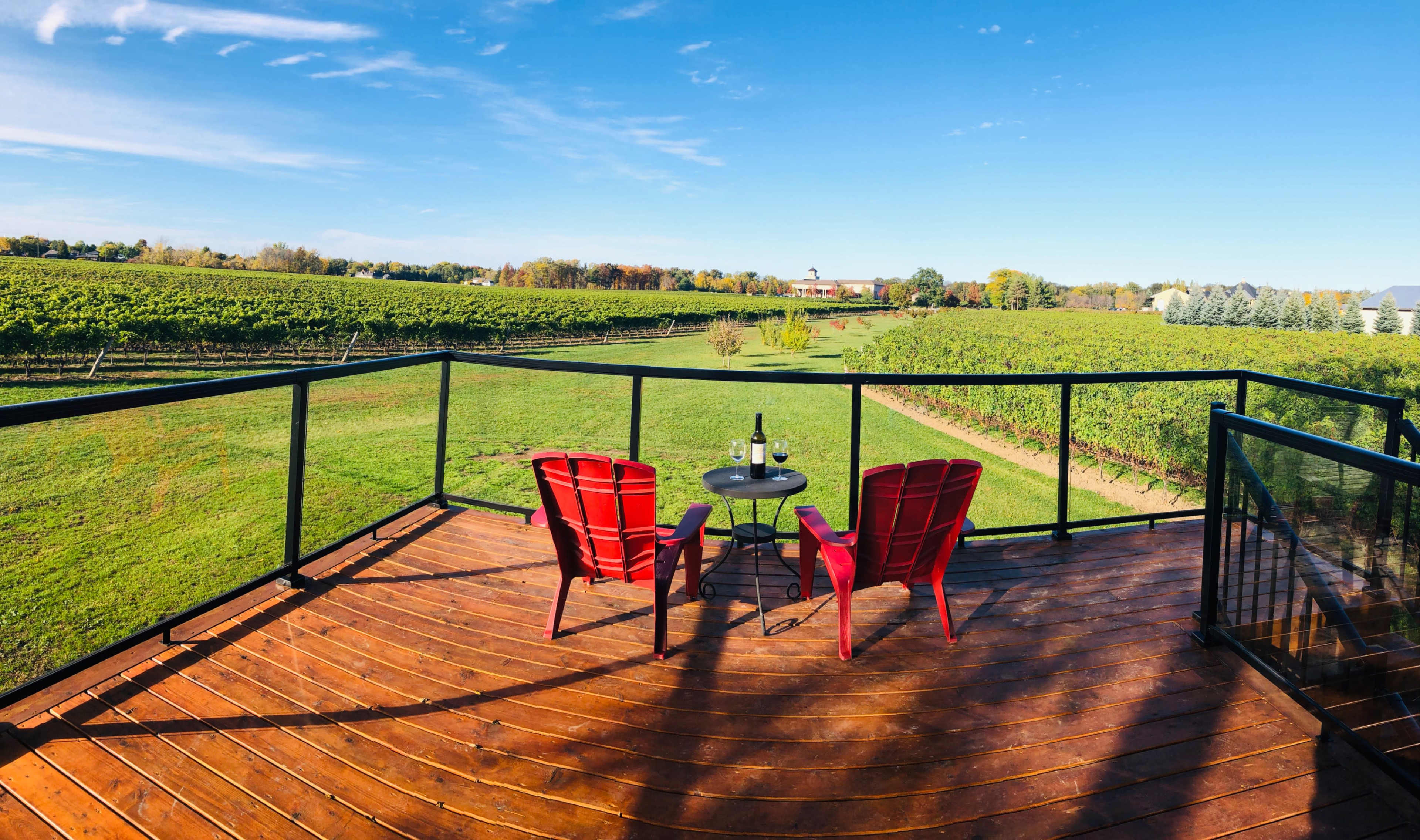 A deck with two red chairs and a small table overlooks a vineyard and a clear blue sky.