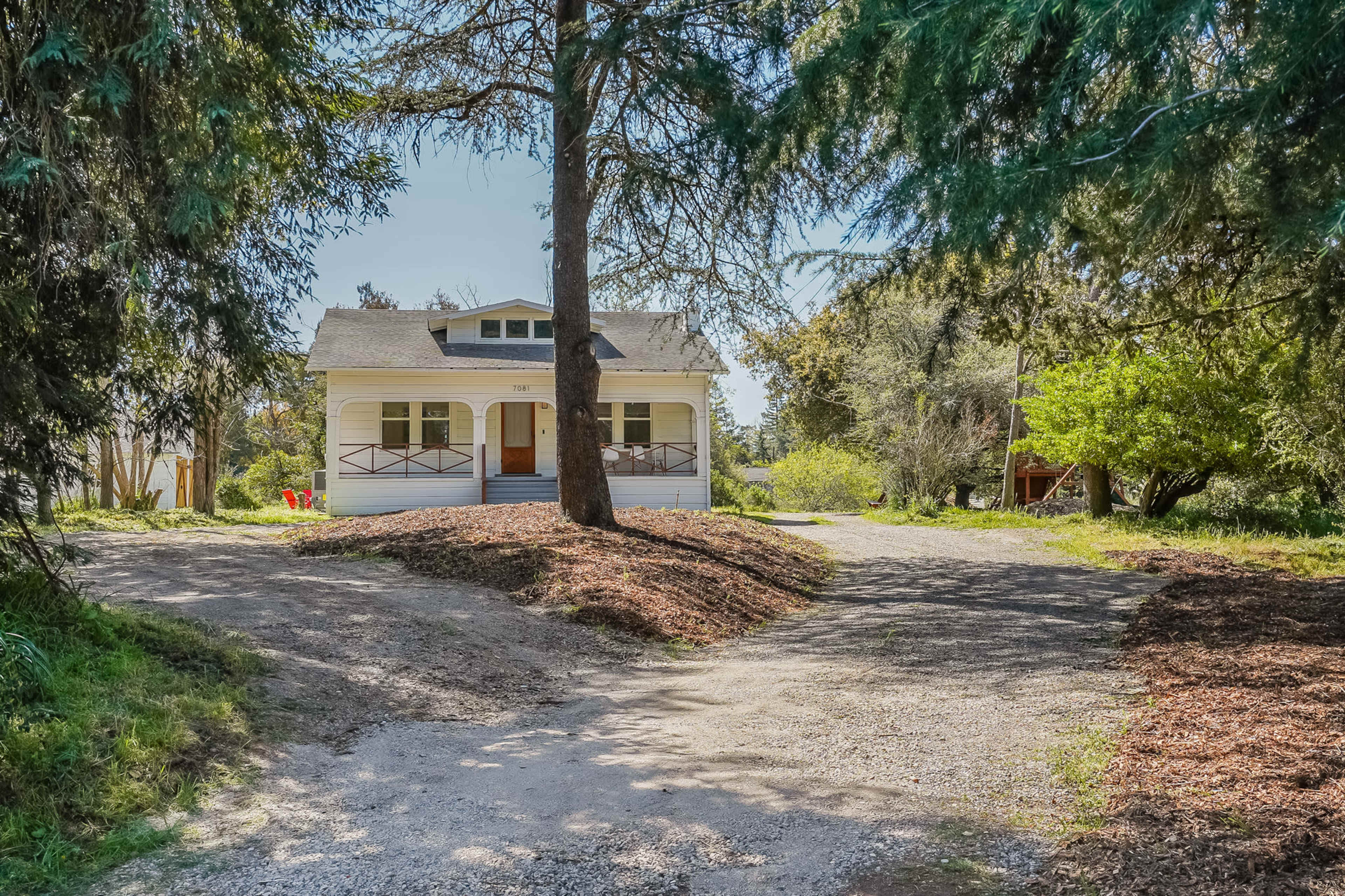 A house with a front porch situated at a fork in a gravel driveway surrounded by trees and shrubs.