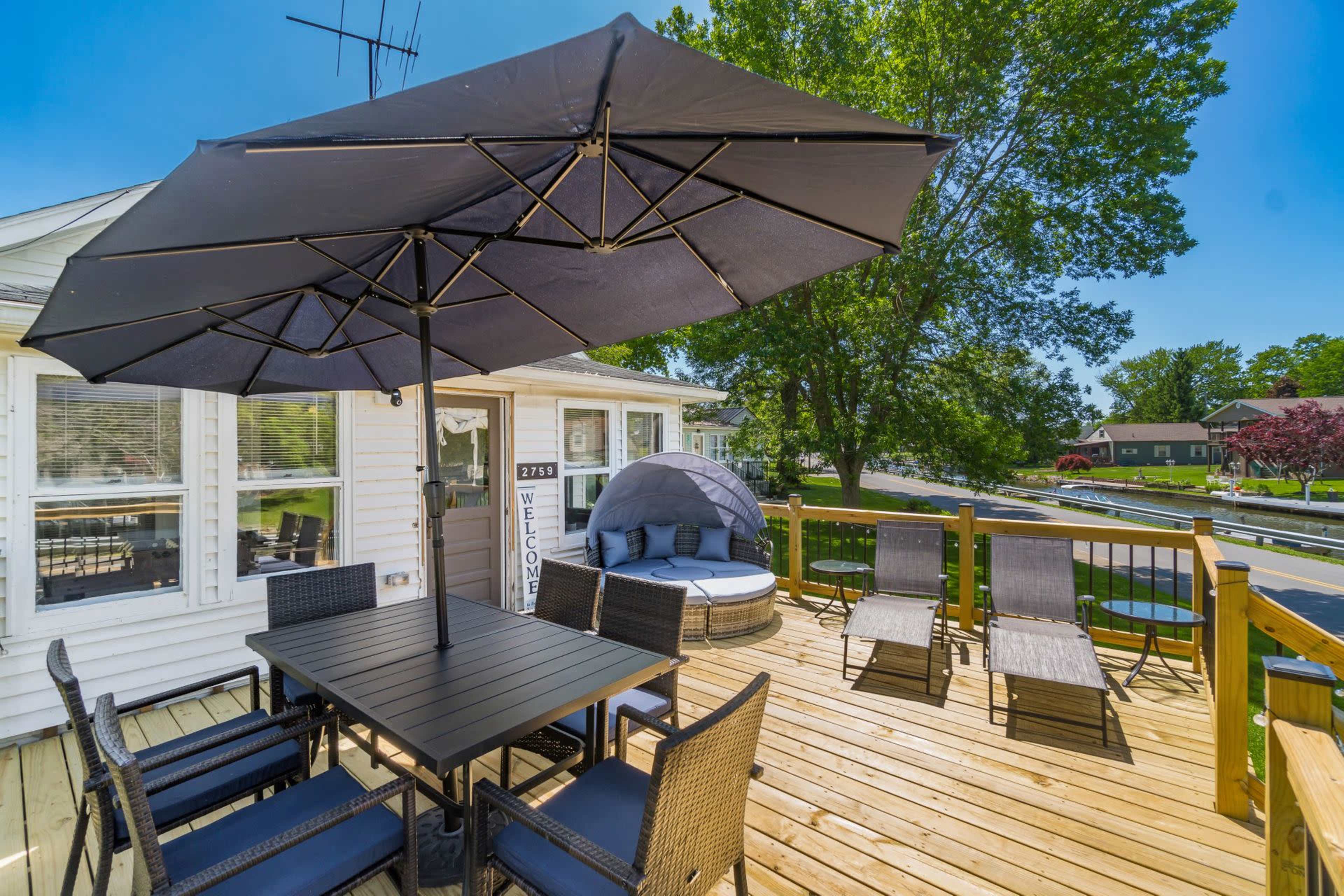A spacious wooden deck features a black umbrella over a table with chairs, surrounded by lounge chairs and views of a neighborhood.