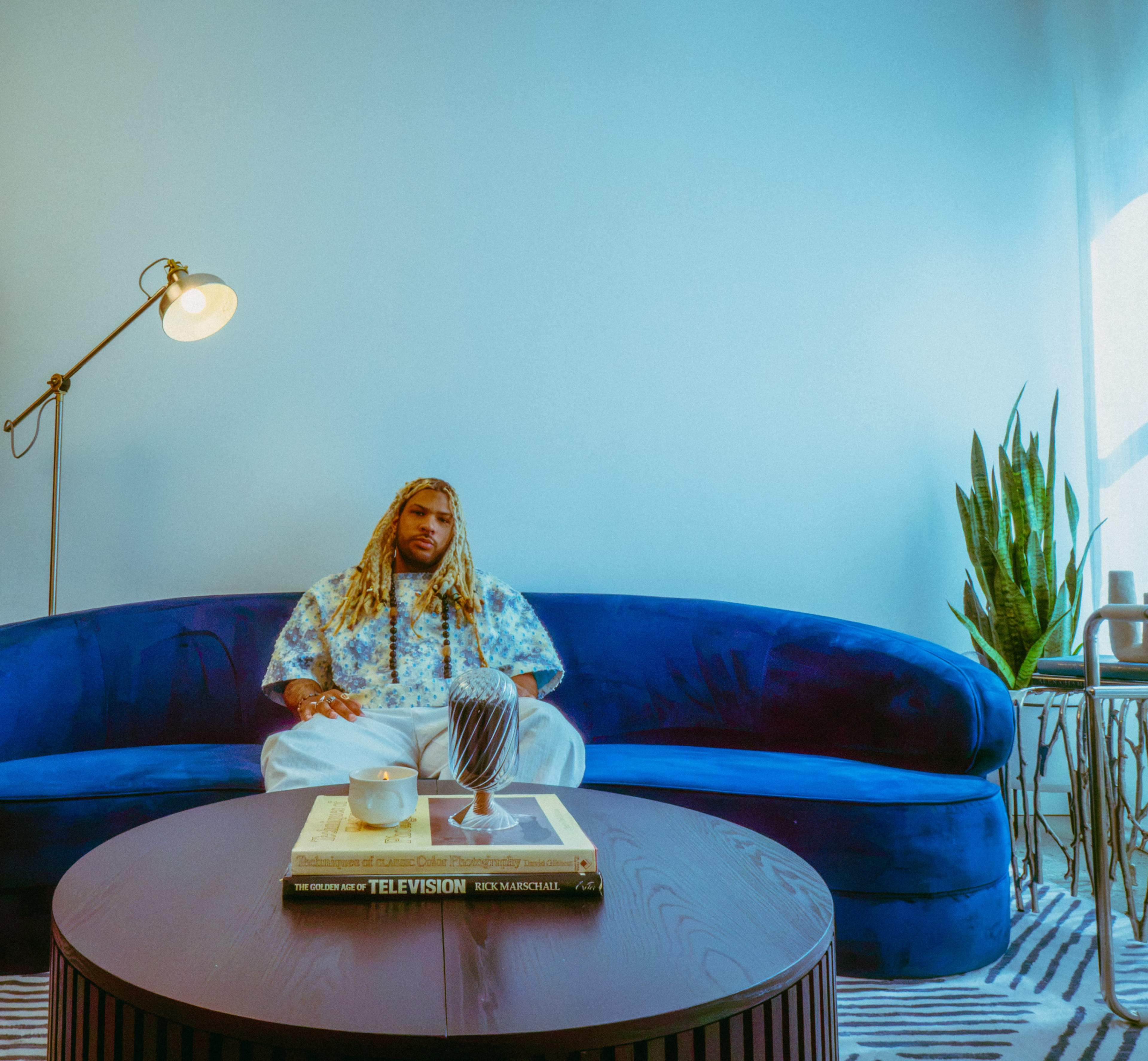 A person with long hair sits on a blue couch in a well-lit room, accompanied by a coffee table featuring books and decorative items.