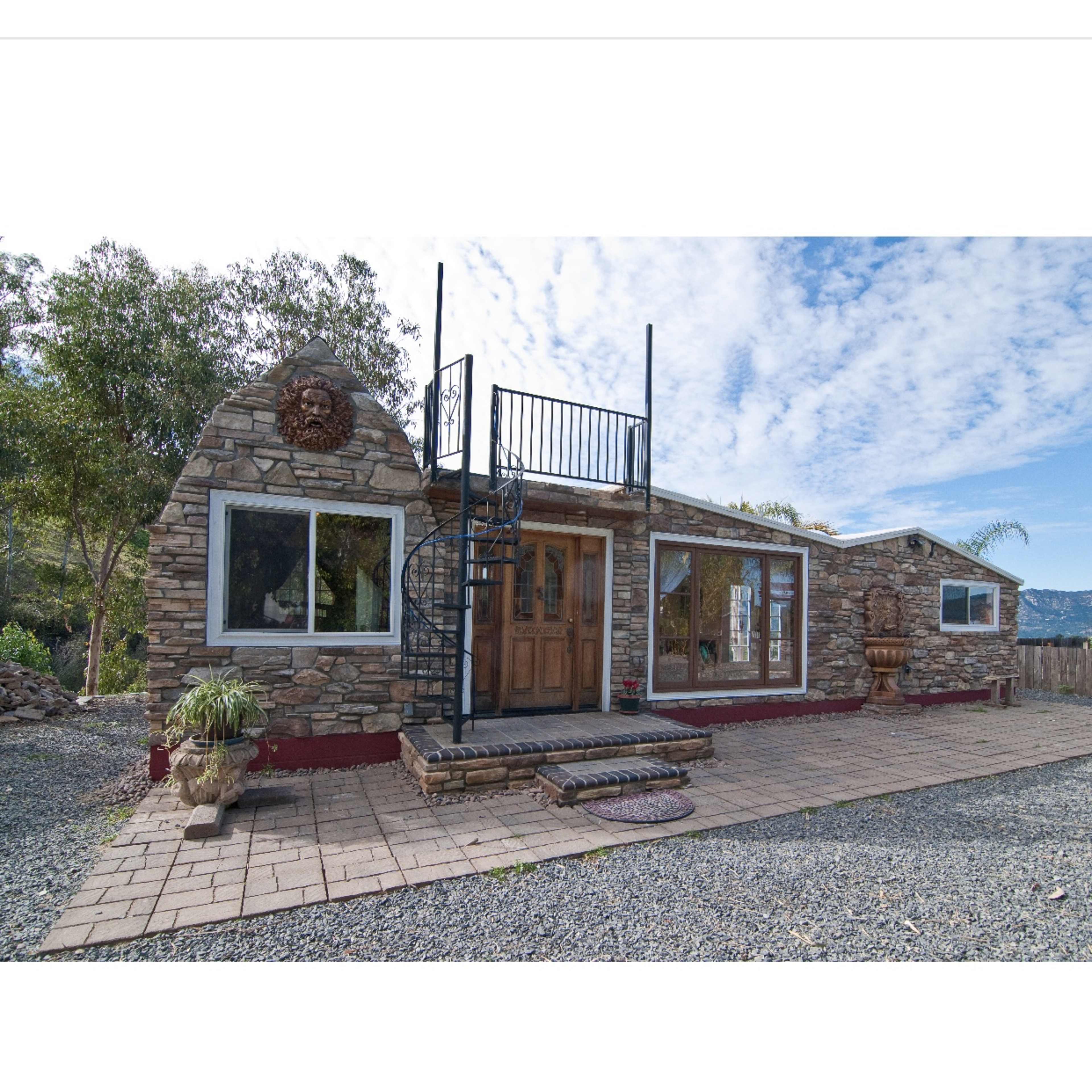 A stone house with large windows and a spiral staircase is set against a cloudy sky.