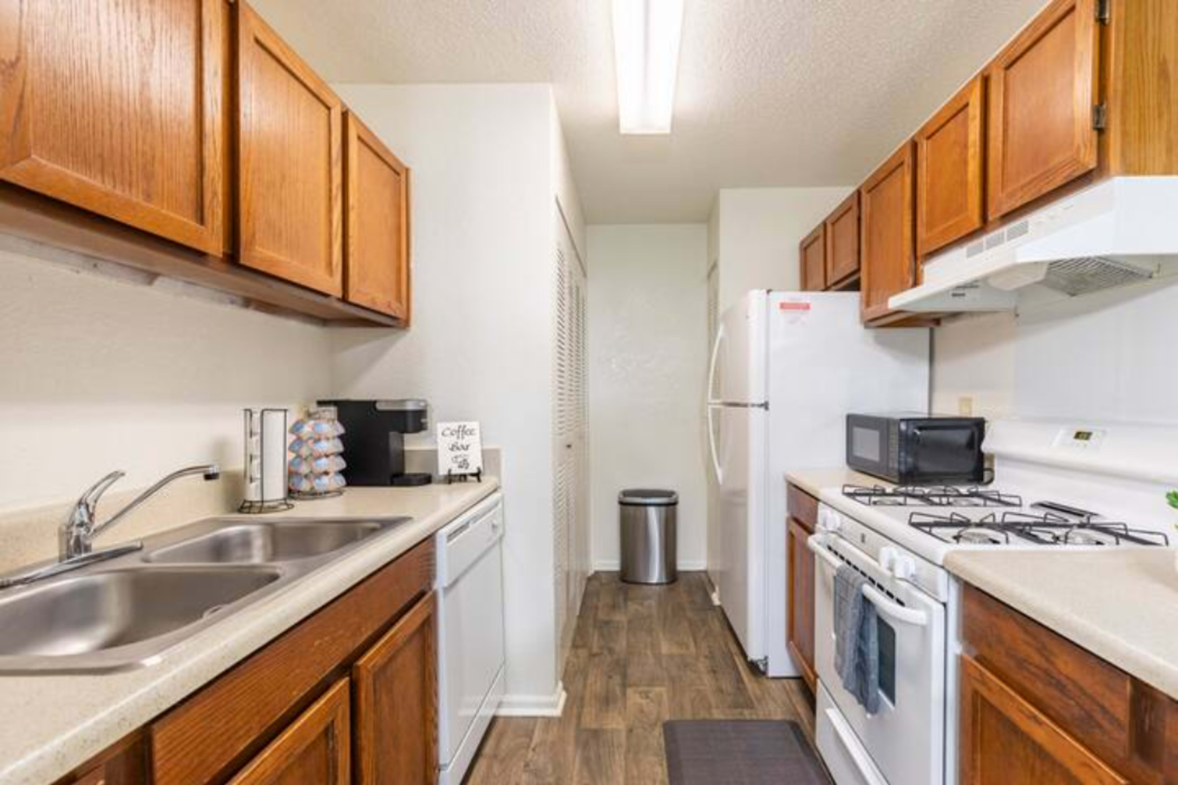 A narrow kitchen features wooden cabinets, a white refrigerator, a stove, and a double sink, with a coffee maker and a small microwave on the counter.