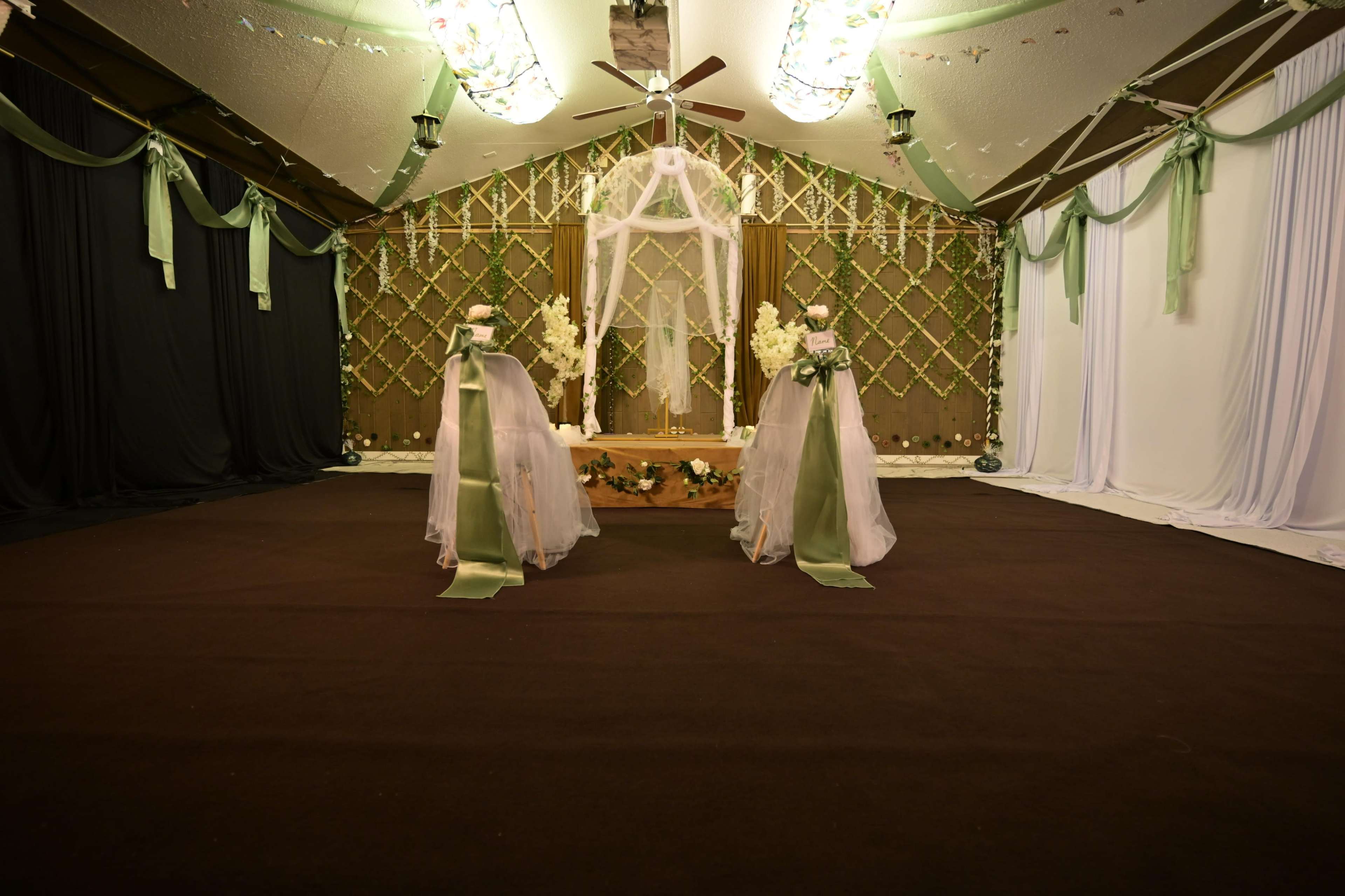 The image shows a decorated indoor venue for a wedding ceremony with a floral altar, draped fabric, and two floral arrangements on stands.