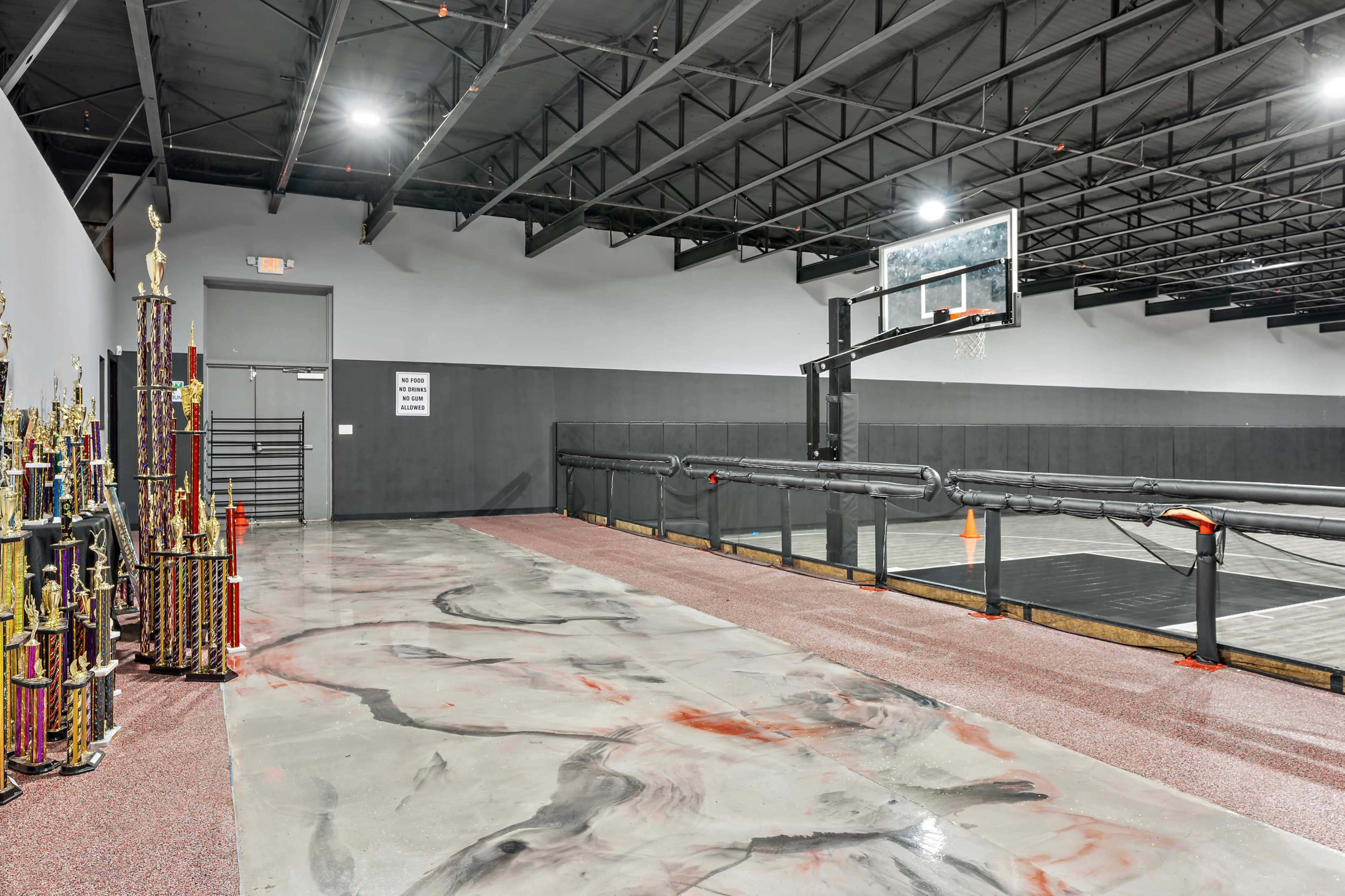 A spacious indoor basketball court with a hoop, surrounded by a trophy display on one side and a polished concrete floor.