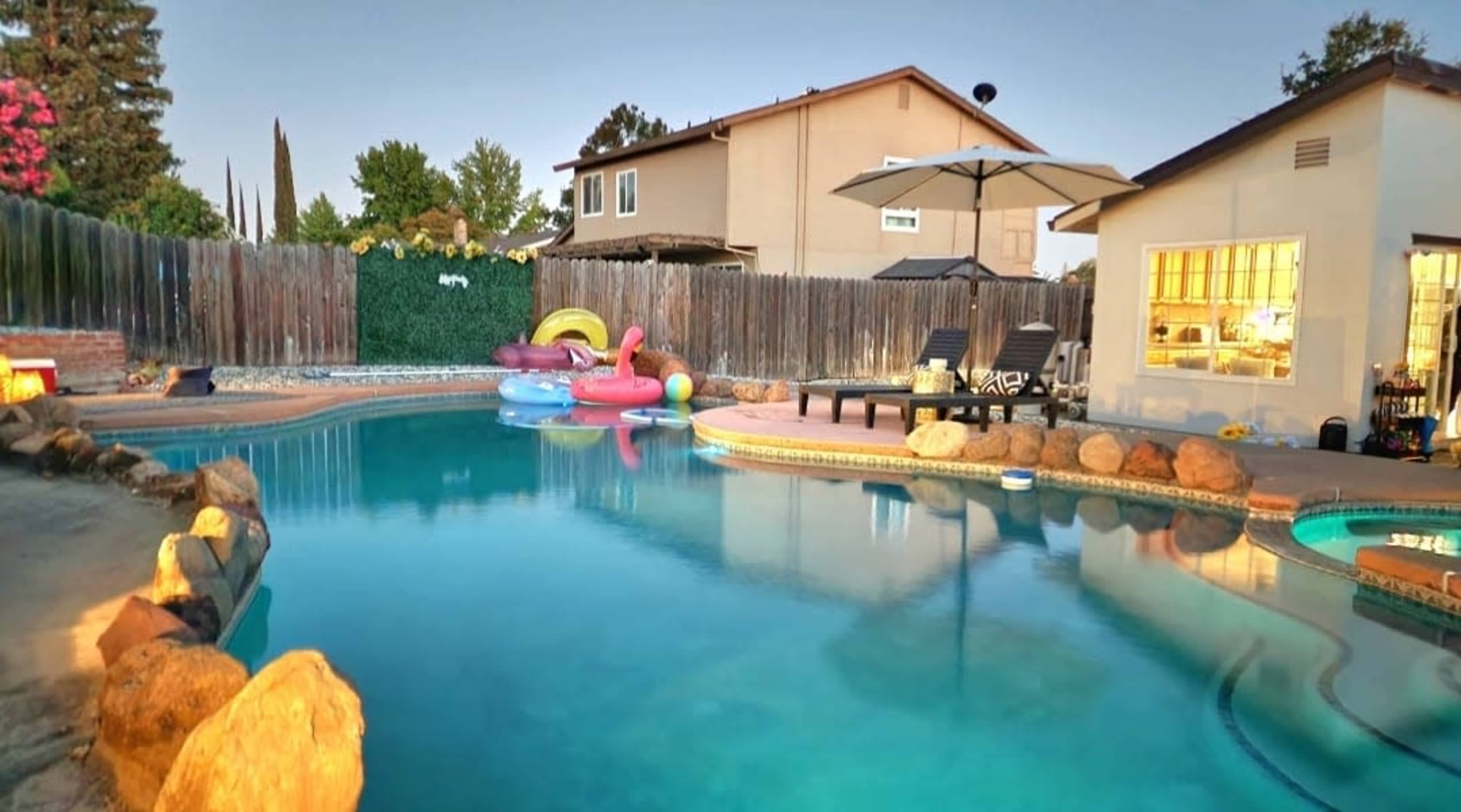 A residential backyard features a swimming pool surrounded by rocks, with inflatable rafts floating in the water and a patio area with chairs and an umbrella.