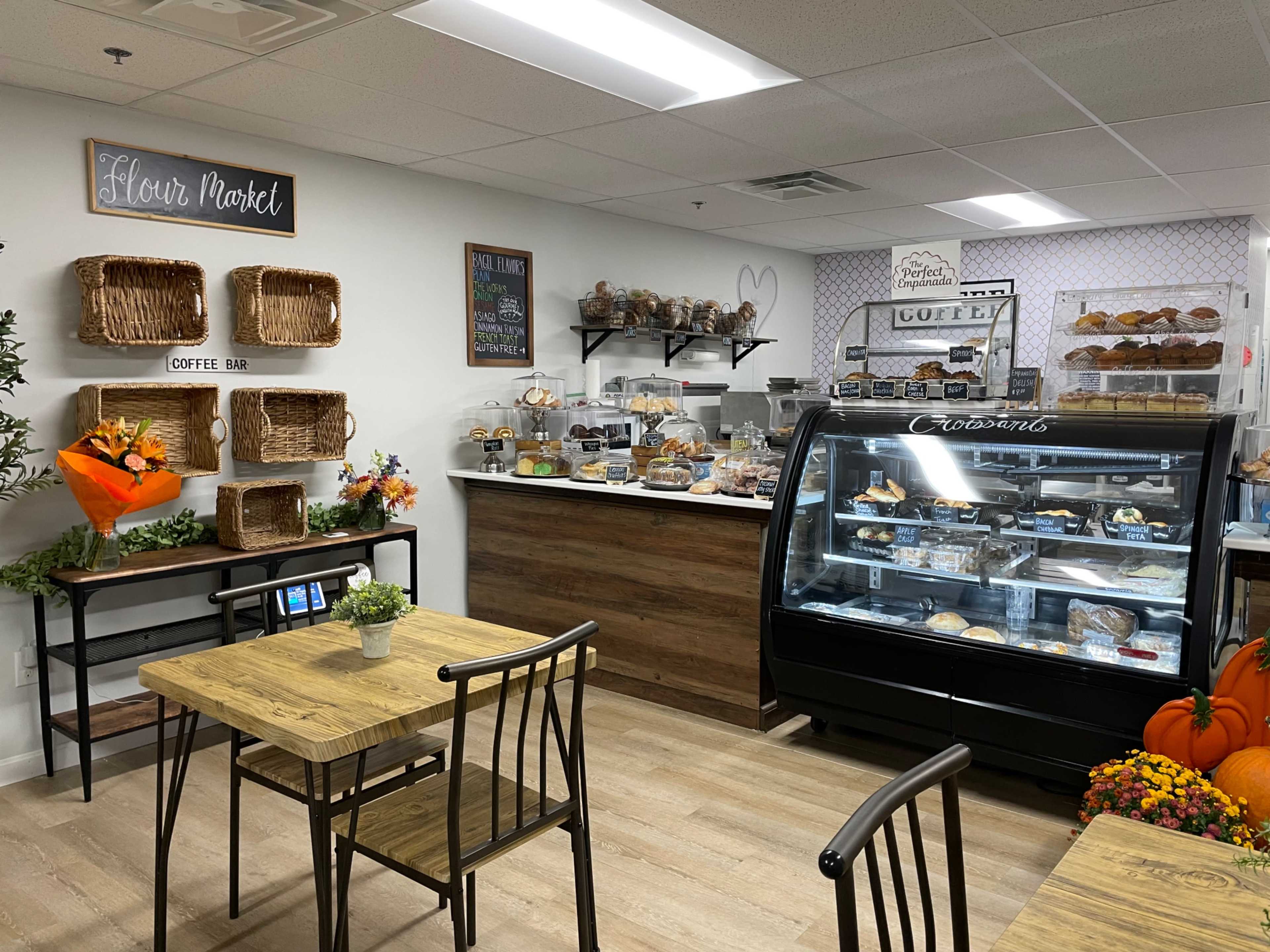 A cozy café interior with a display case of baked goods, shelves of pastries, and a small seating area featuring wooden tables and chairs.