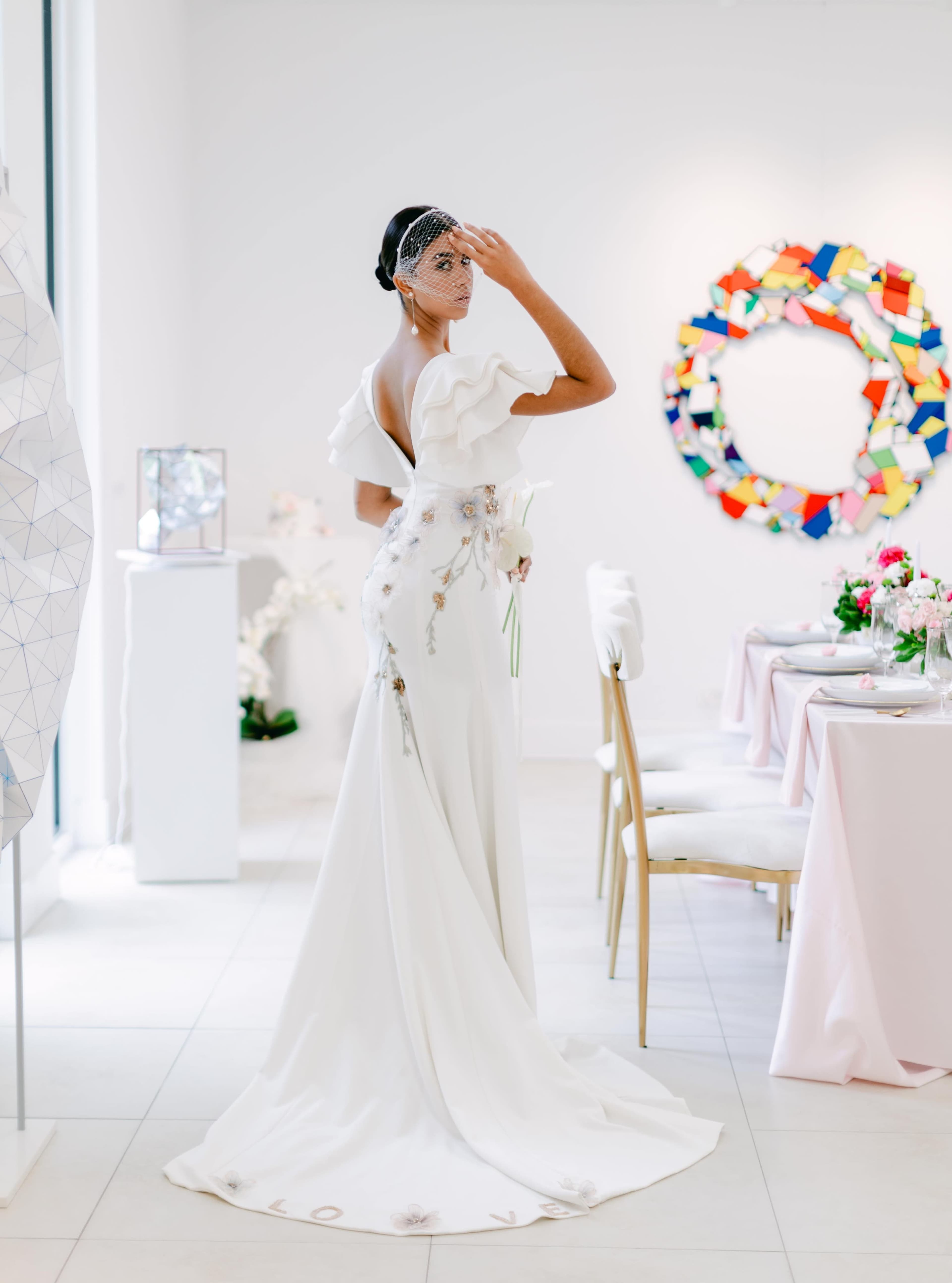 A woman in a white dress with ruffled sleeves poses with her back turned in a modern, elegantly decorated room featuring art and a dining setup.