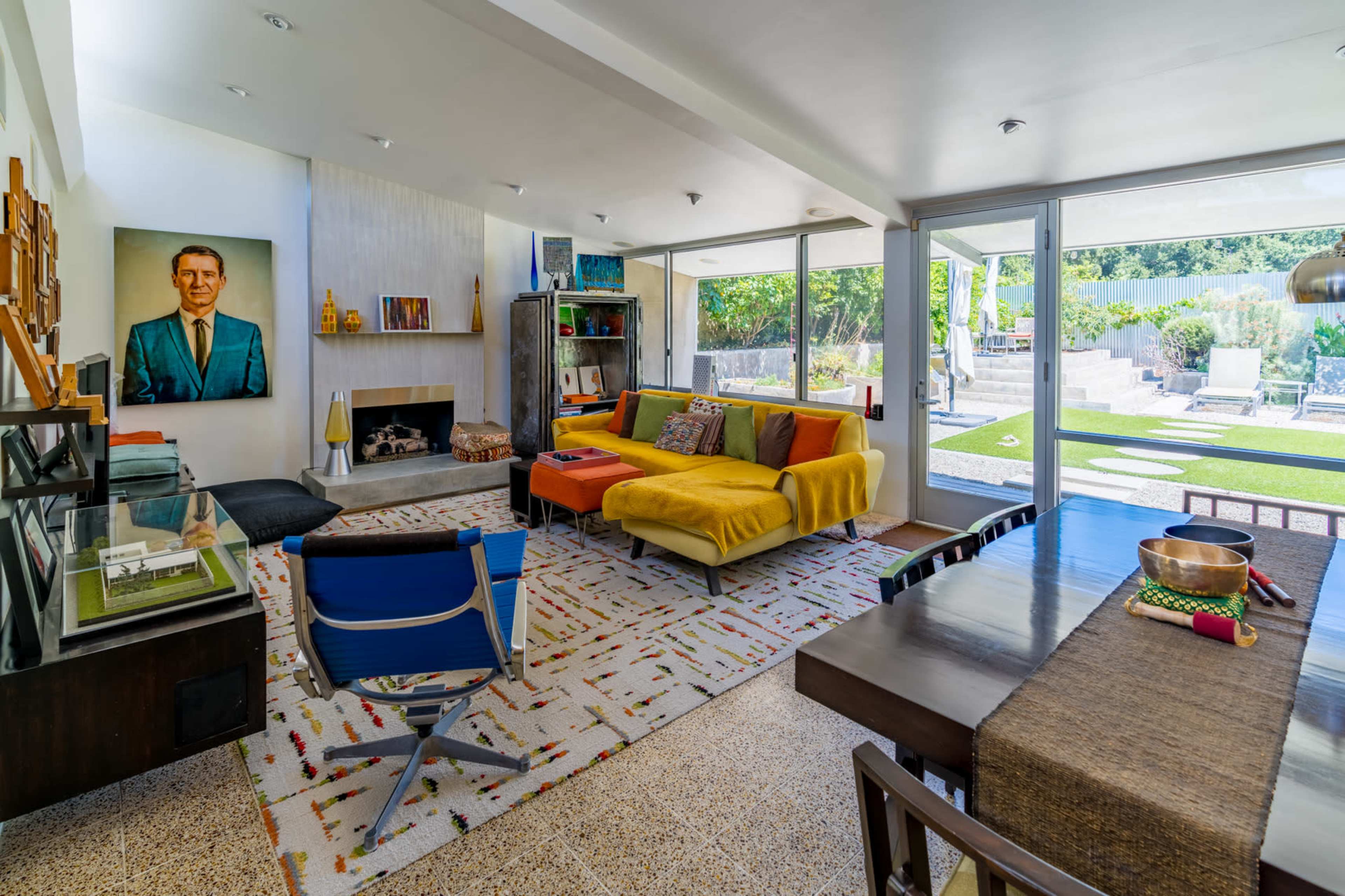 A modern living room with a colorful sectional sofa, a fireplace, a dining table, and large windows overlooking a garden area.