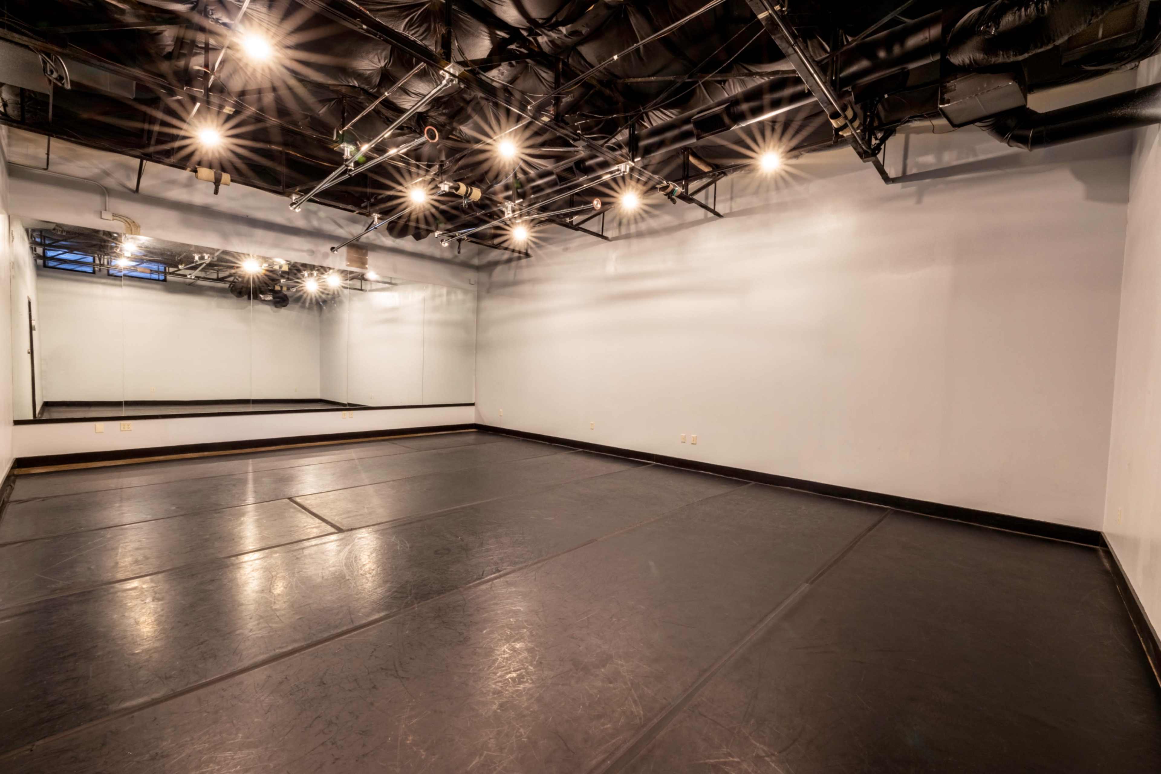 The image shows an empty dance studio with a mirrored wall and black flooring, illuminated by overhead lights.