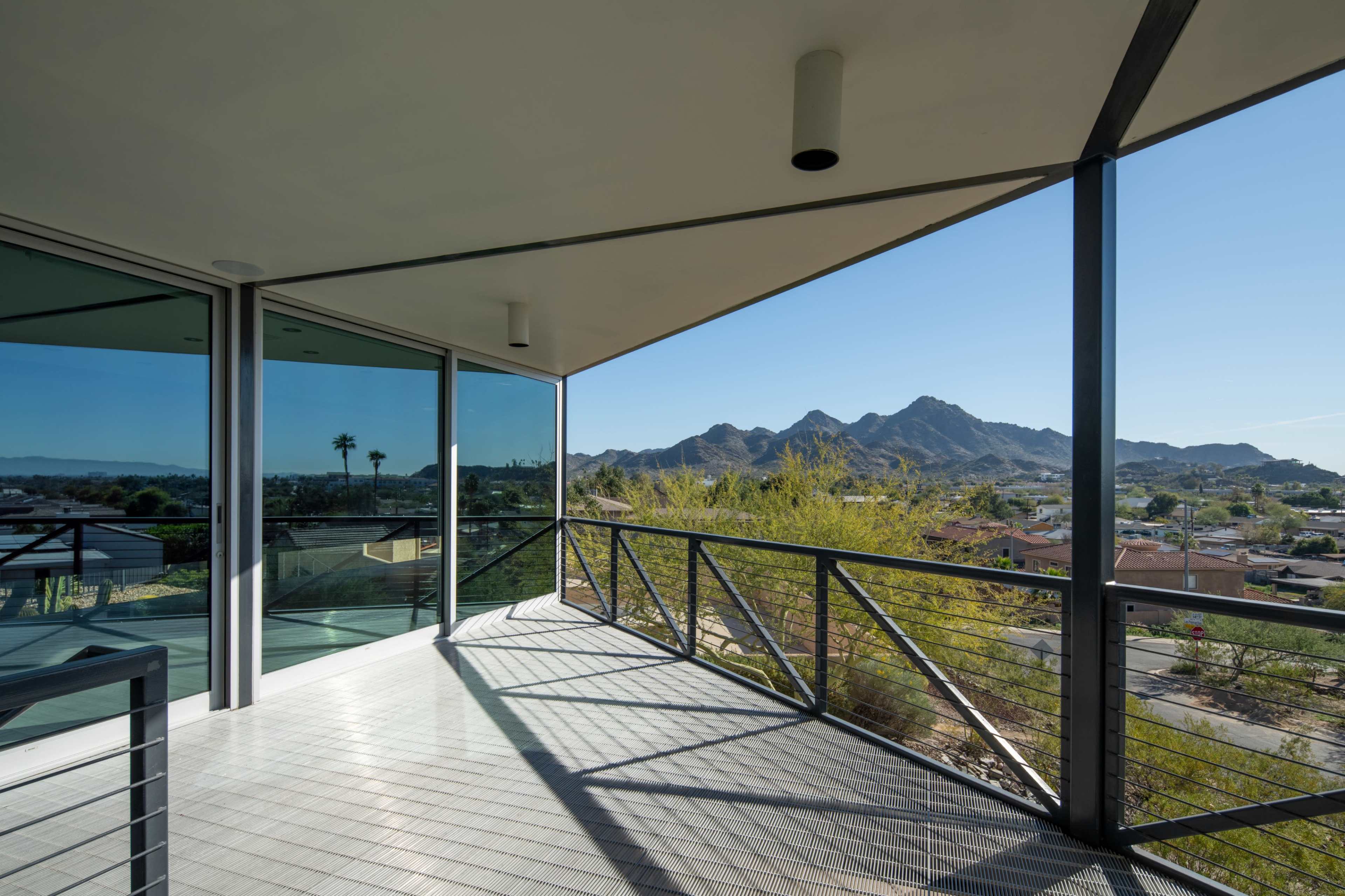 The image shows a modern balcony with a metal railing overlooking a mountainous landscape under a clear blue sky.