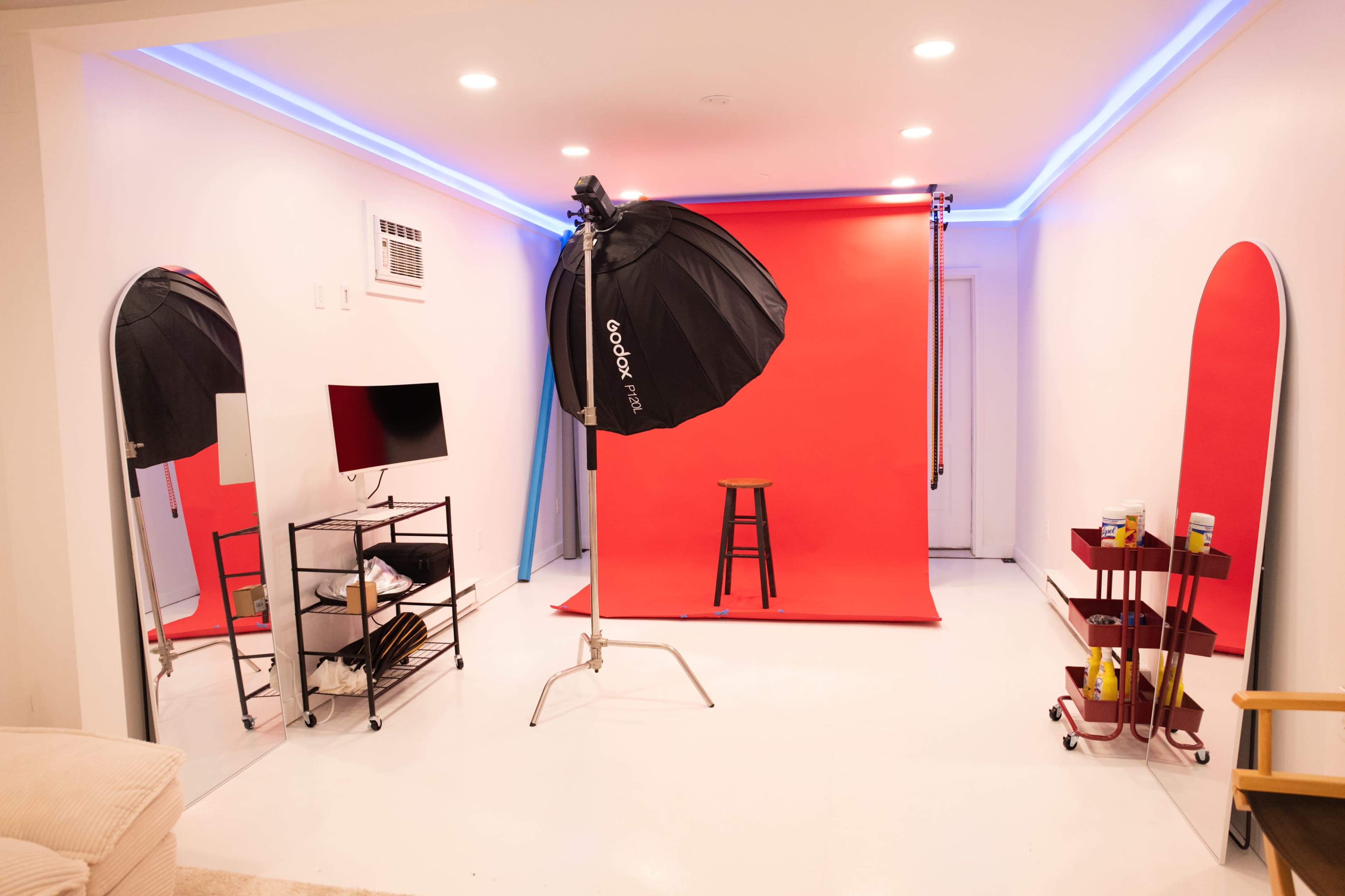 A photography studio setup features a red backdrop, a stool, softbox lighting, and various props on shelves.