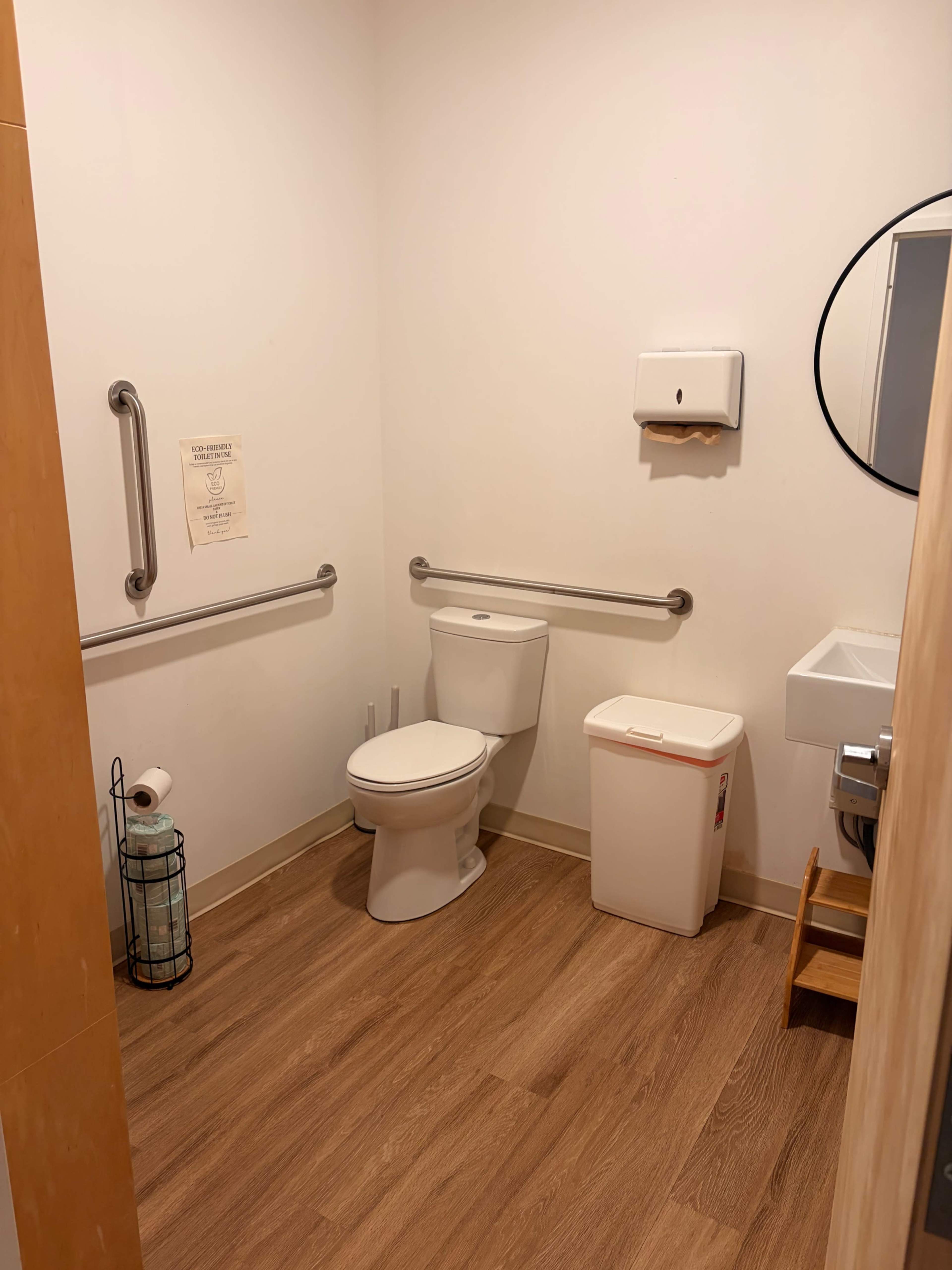 The image shows a small restroom with a toilet, a handwashing sink, a waste bin, and grab bars on the walls.