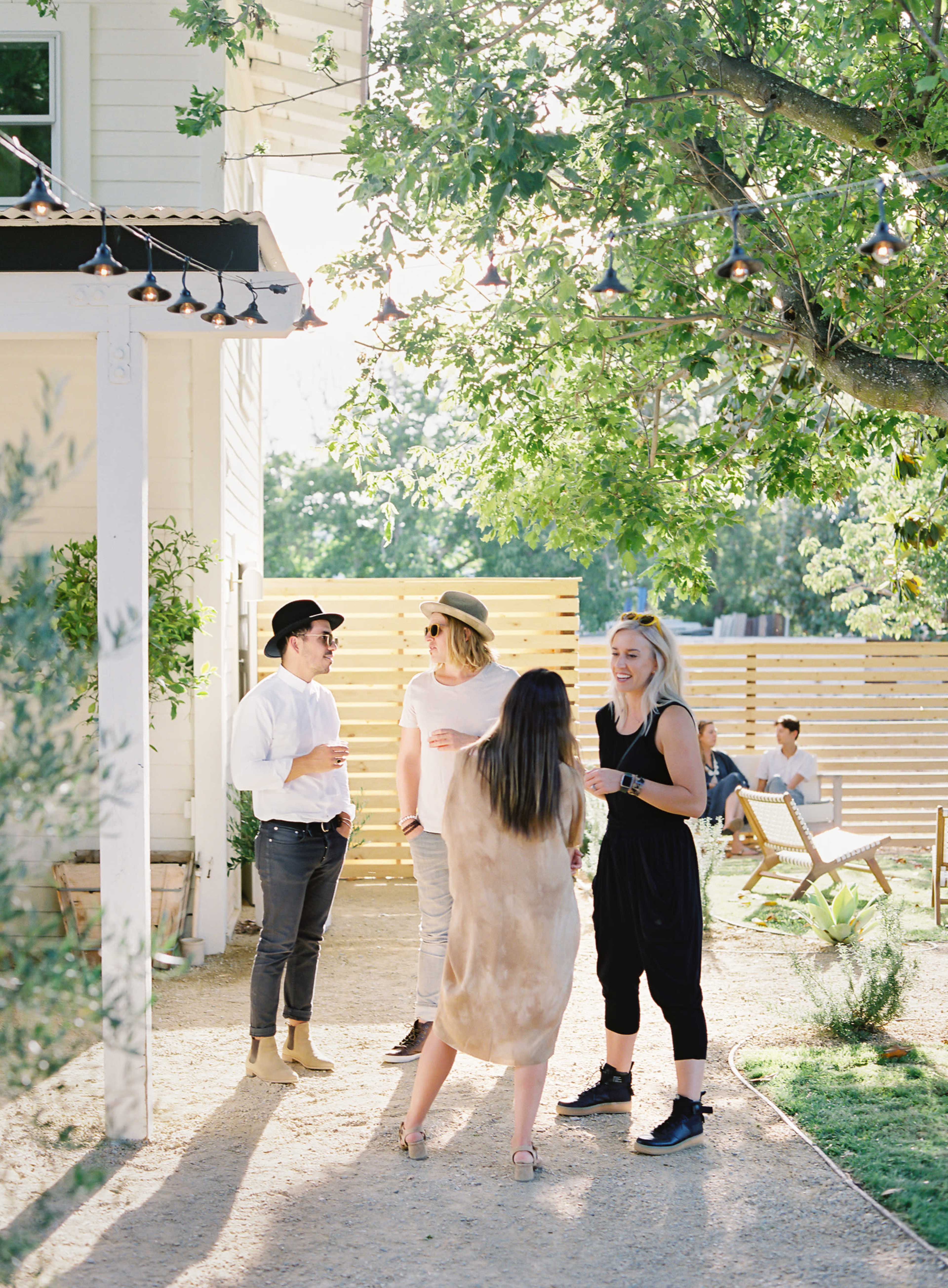 A group of four people stands in a sunlit outdoor area, engaged in conversation near a building and decorative string lights.