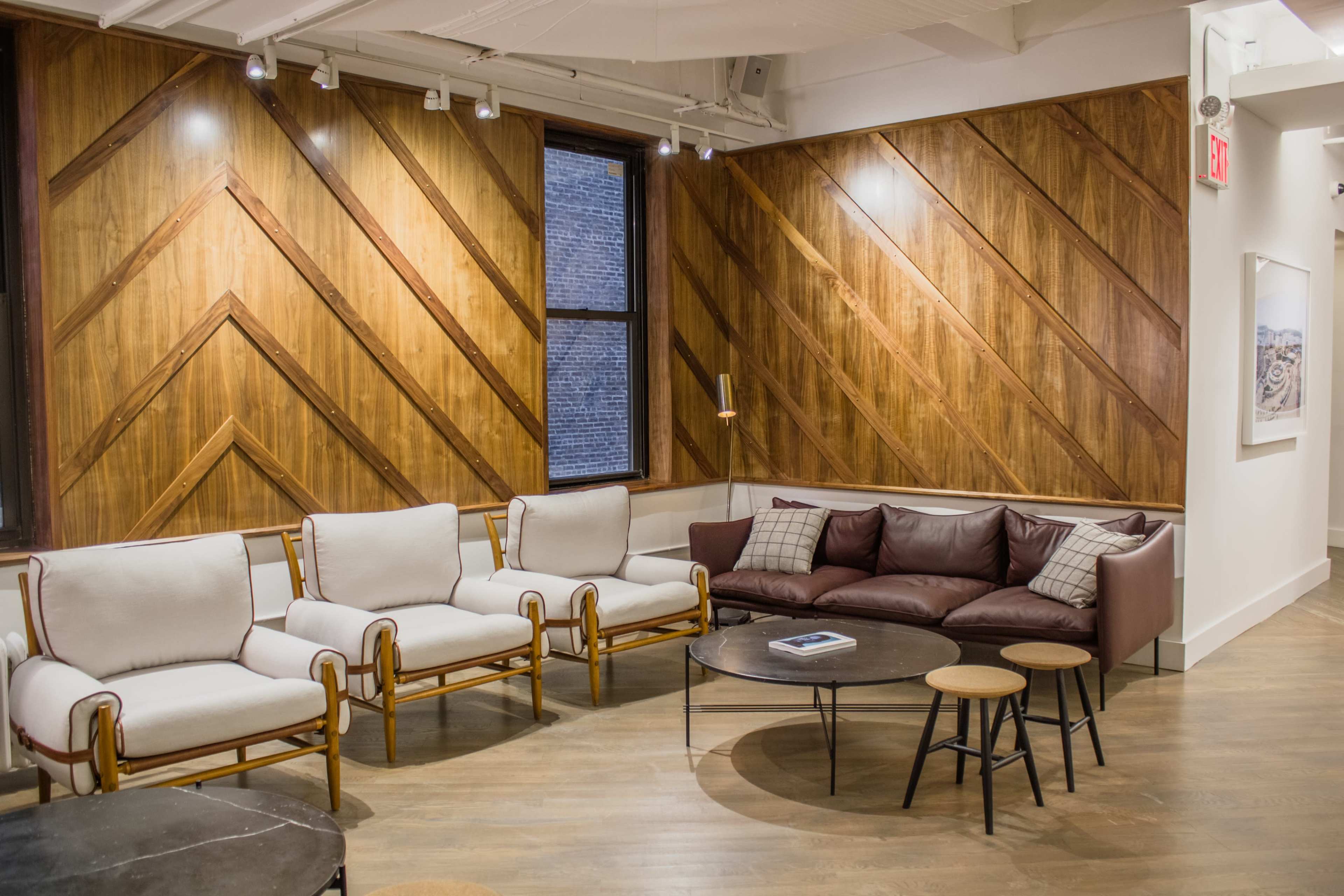 The image shows a cozy seating area with a brown leather sofa, beige chairs, and a round table set against a wall with a geometric wooden design.