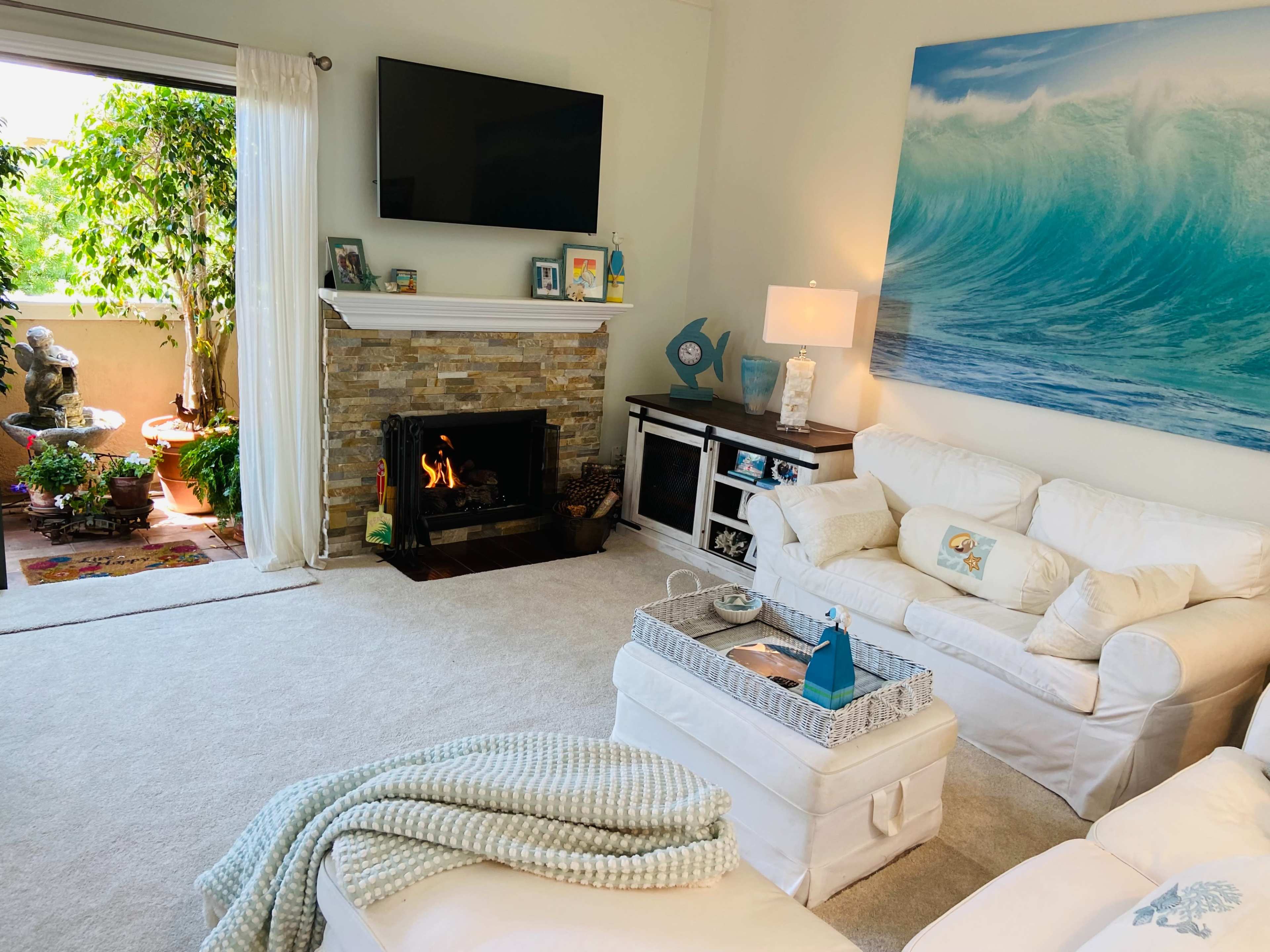 A cozy living room featuring a white sectional sofa, a stone fireplace, a large television mounted above the mantel, and a view of an outdoor area with greenery.
