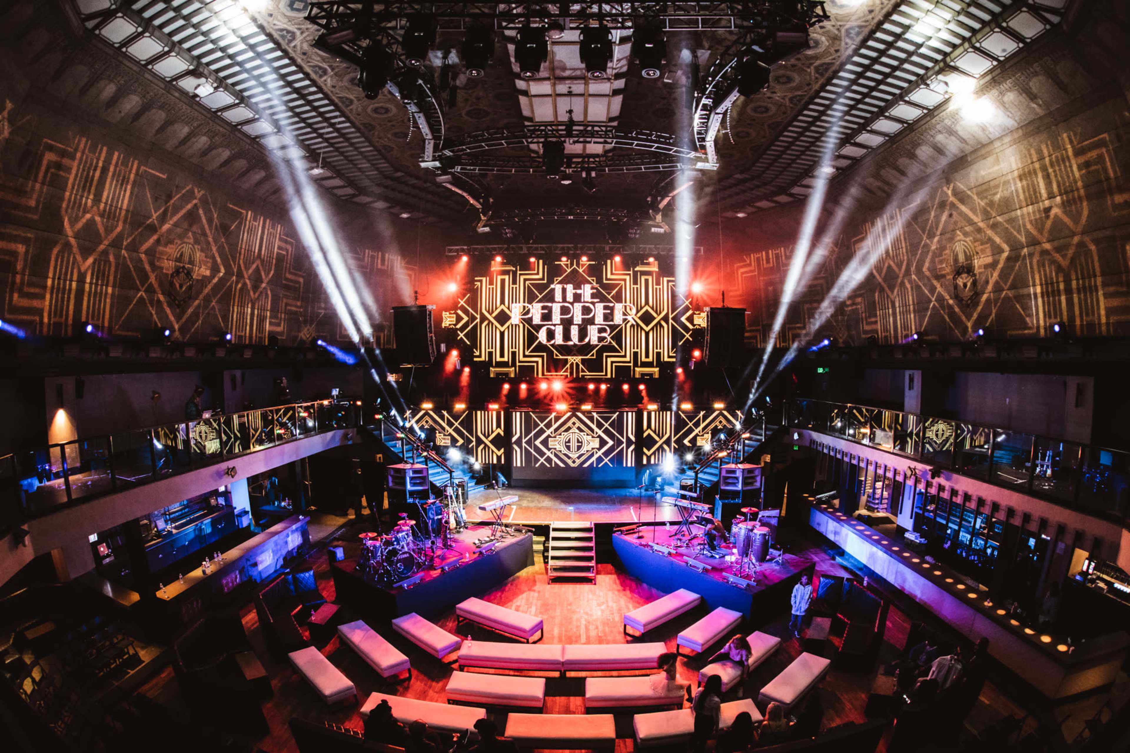 An illuminated nightclub interior featuring a stage, various musical instruments, and seating areas, with vibrant lighting and a large screen displaying "The Pepper Club."