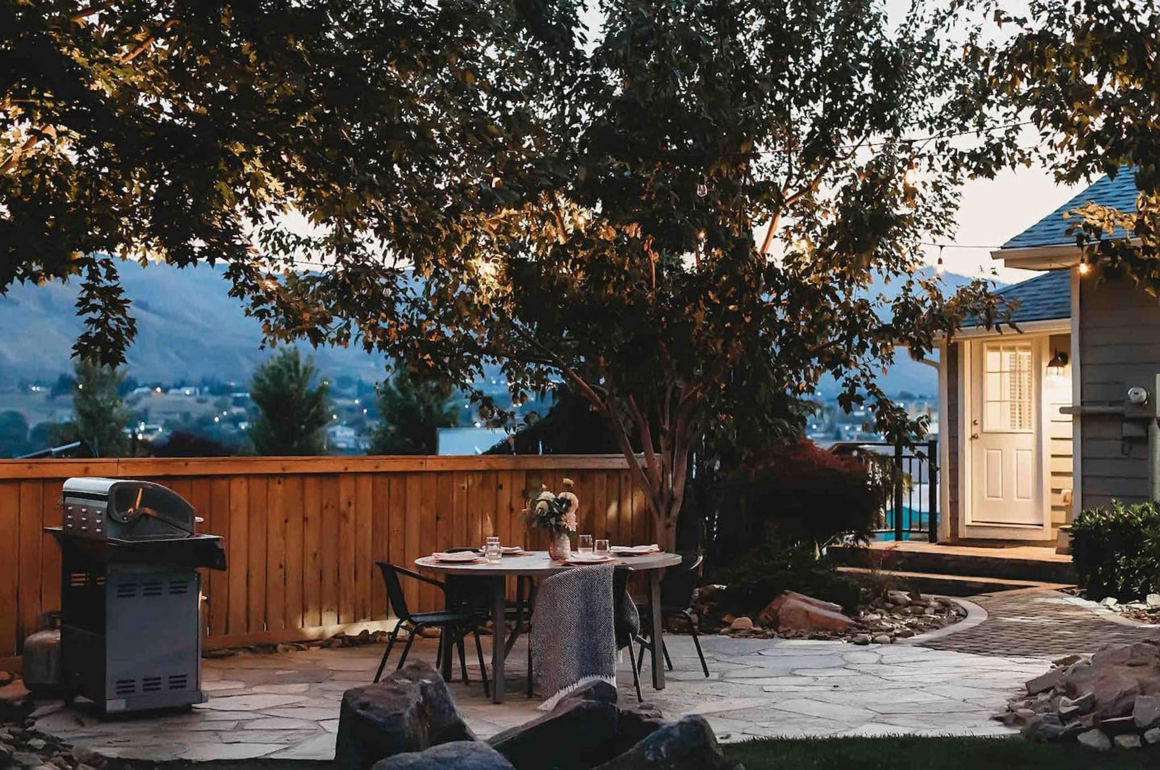 A patio area features a table set for dining, a grill, and a pathway leading to a house, surrounded by trees and rocks.