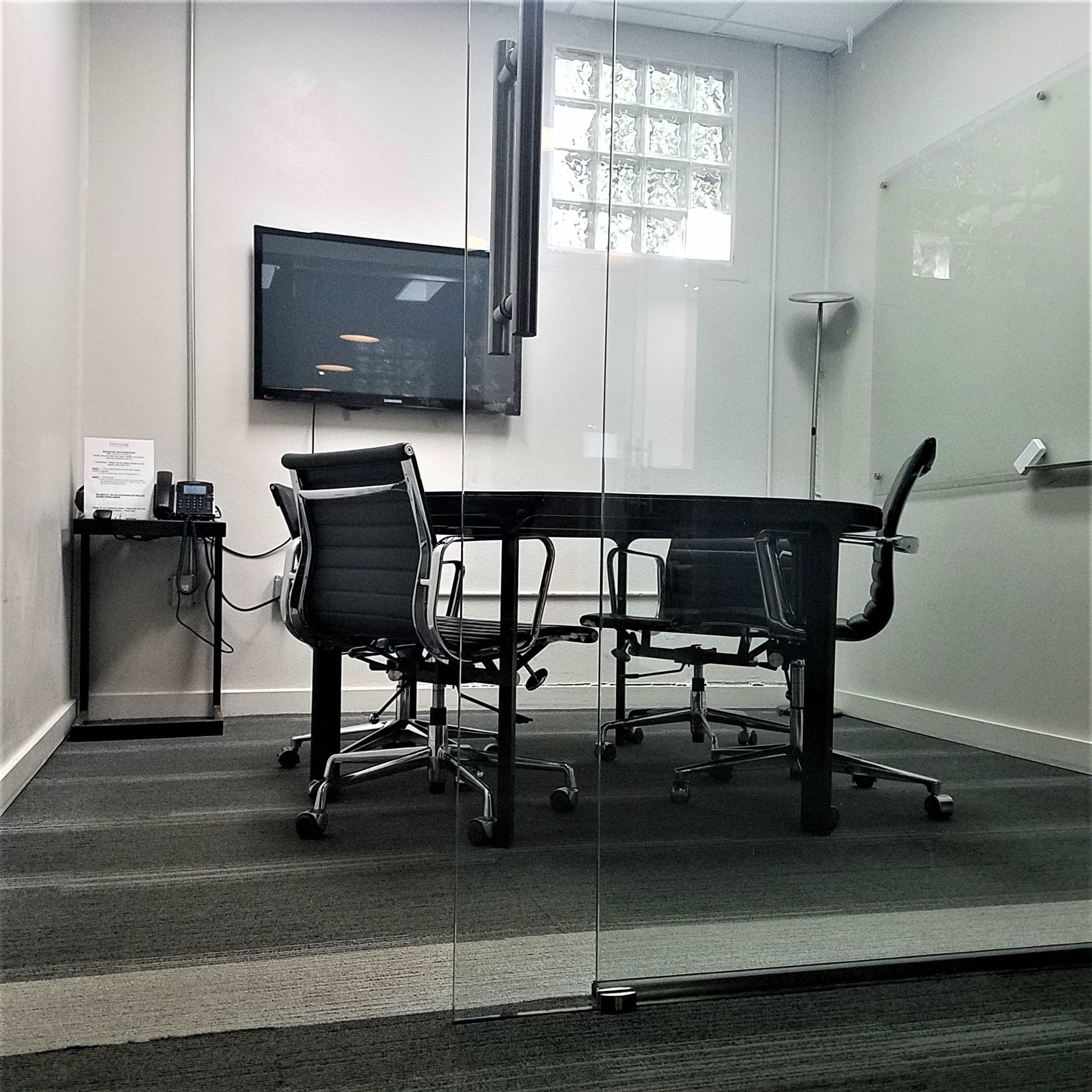 The image shows a conference room with a glass wall, a large black table, four chairs, a television mounted on the wall, and a phone on a side table.