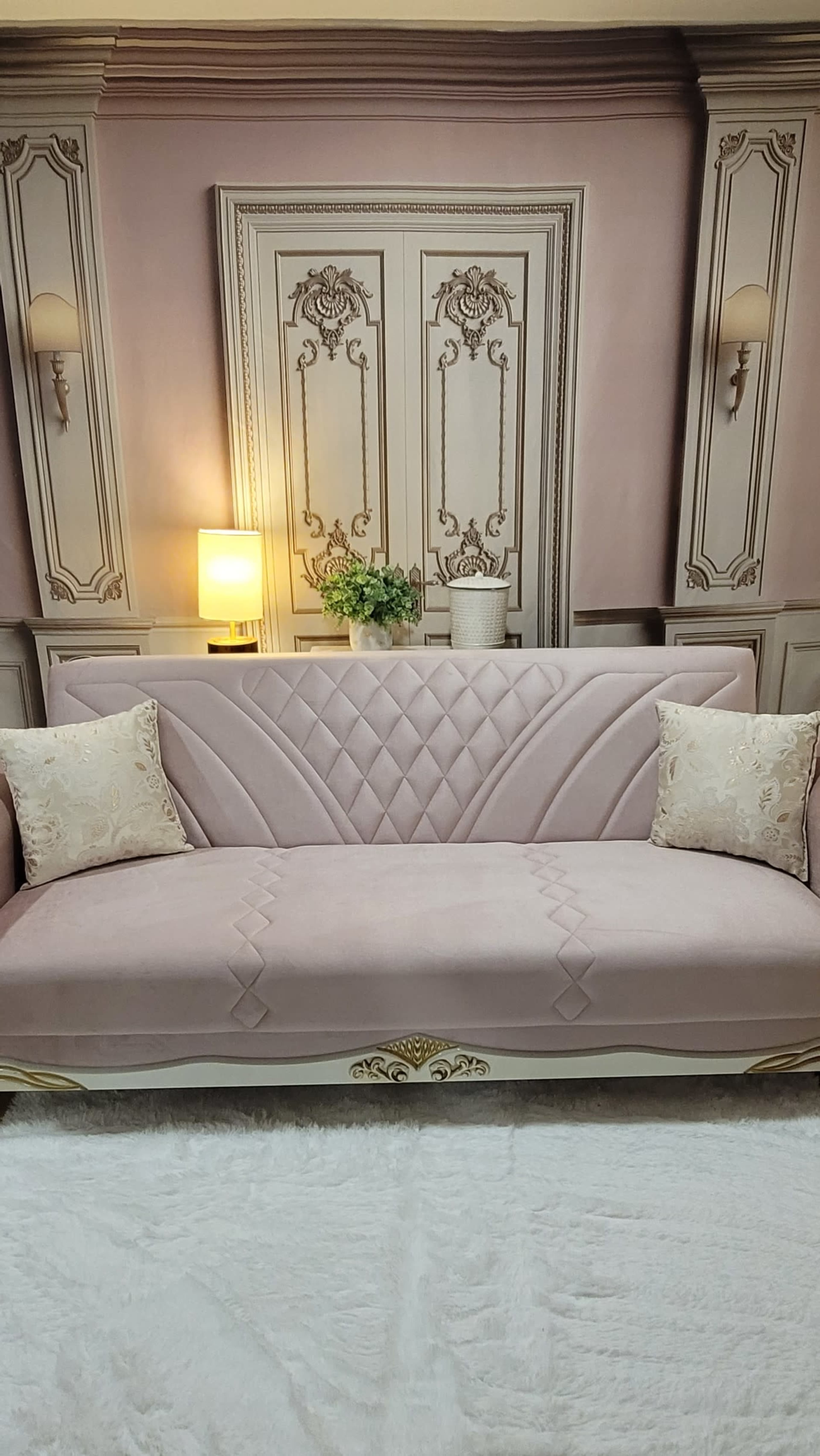 The image features a pink sofa with decorative pillows, set against a softly colored wall with ornate paneling and a lamp in the background.