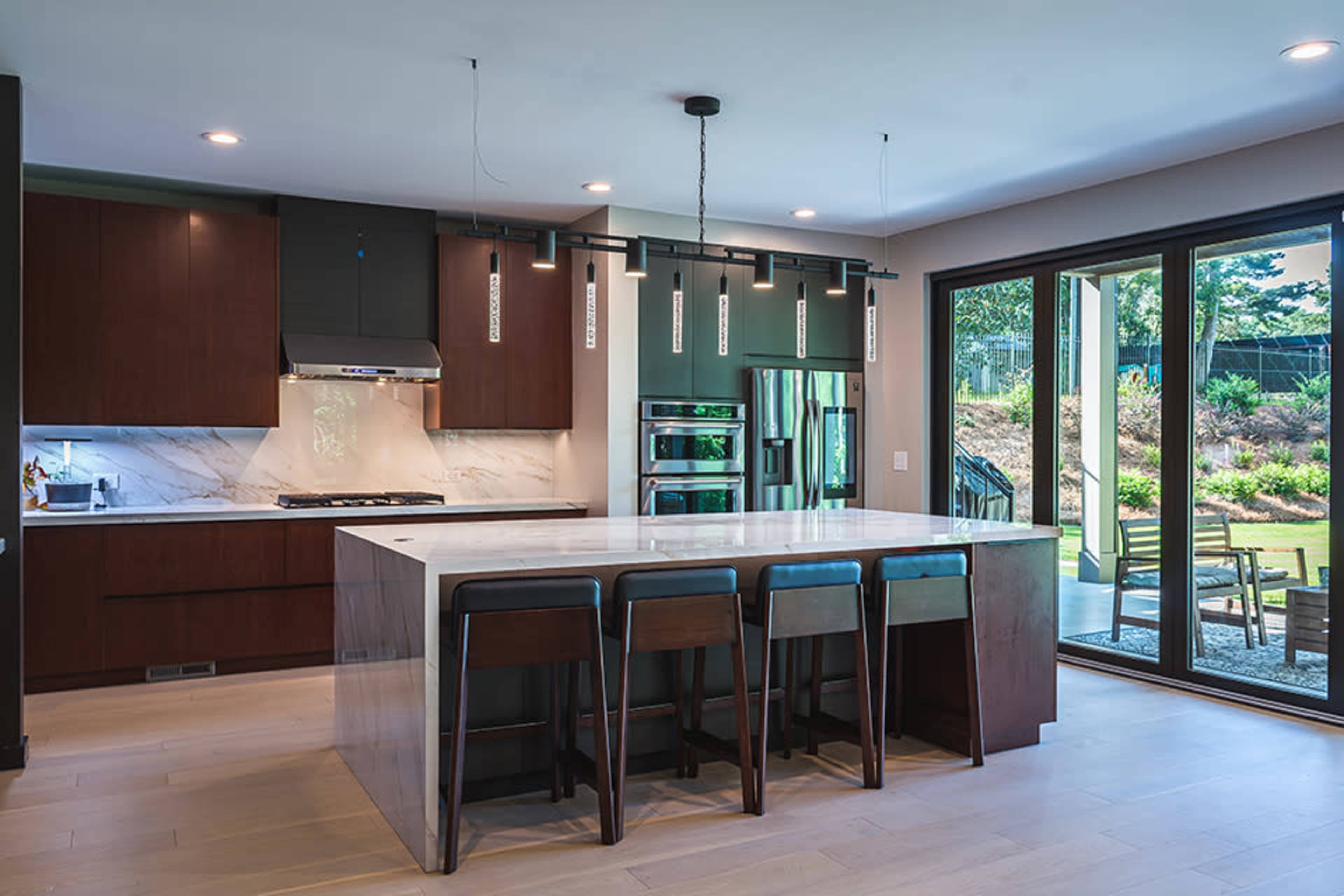 A modern kitchen features a large island with bar stools, dark cabinetry, and sliding glass doors leading to an outdoor patio.