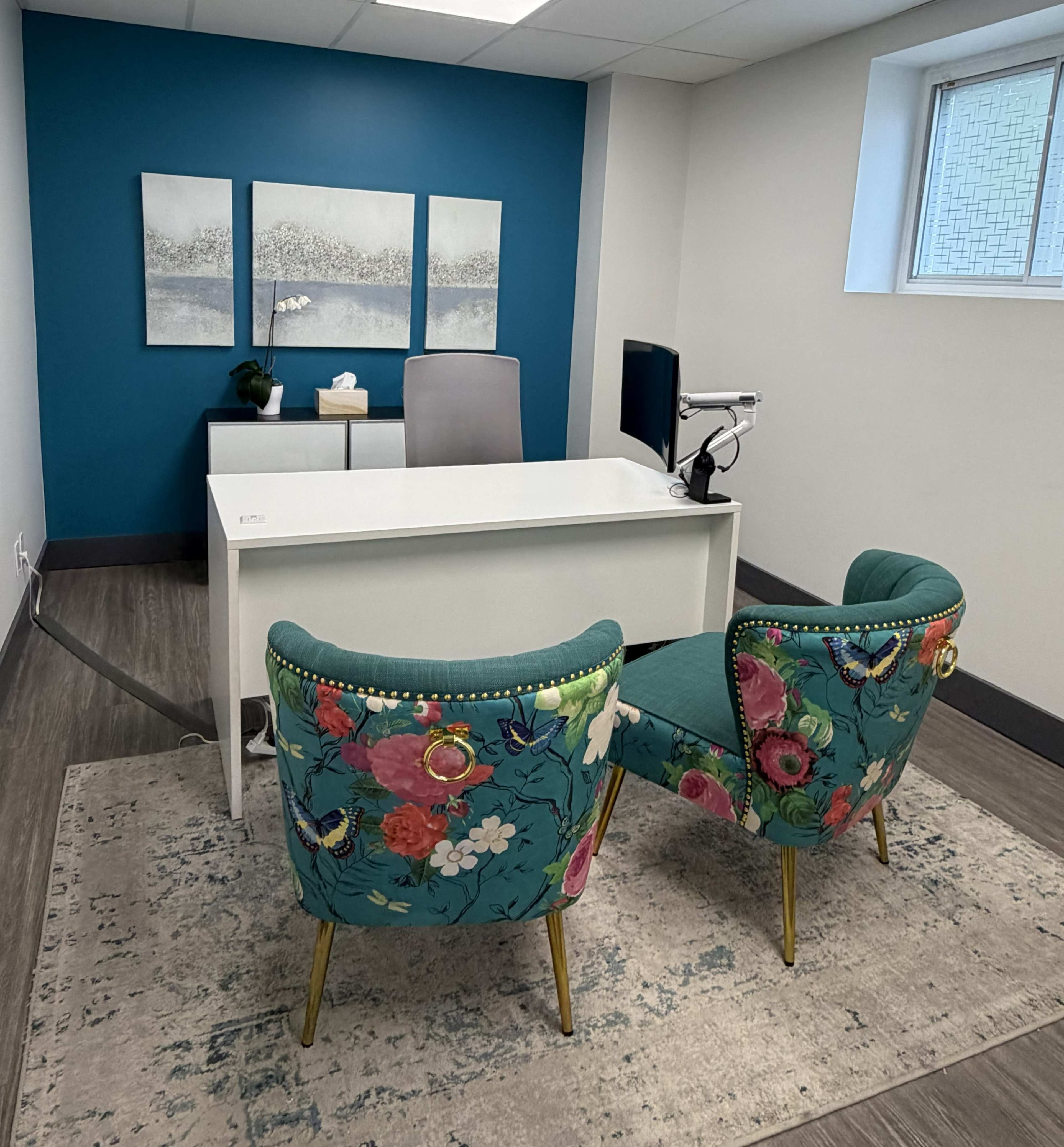 The image shows a modern office space featuring a white desk, a computer, and two floral-patterned chairs placed on a rug.