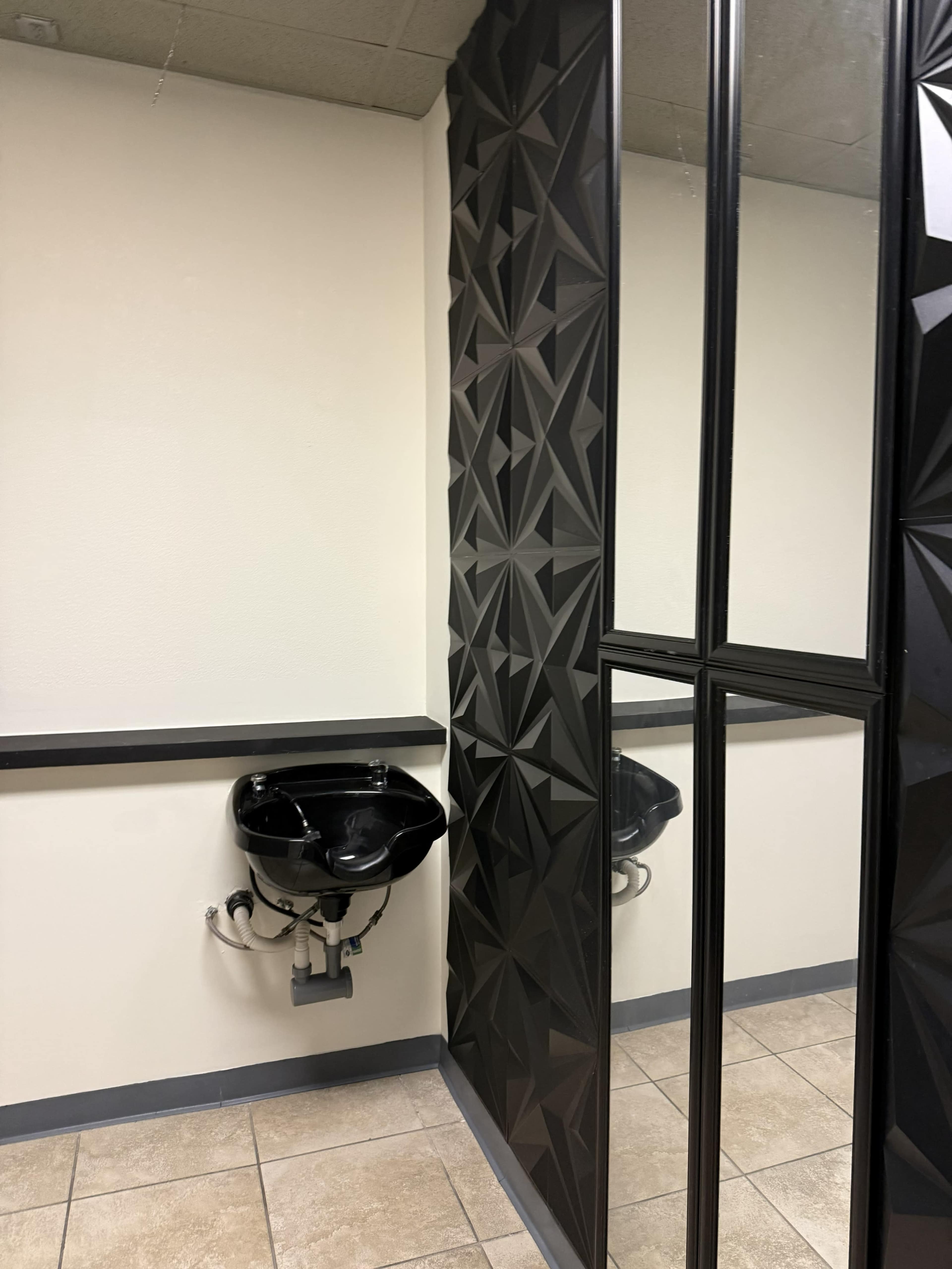 The image shows a corner of a restroom featuring a black sink mounted on the wall, a textured black panel, and large mirrors.