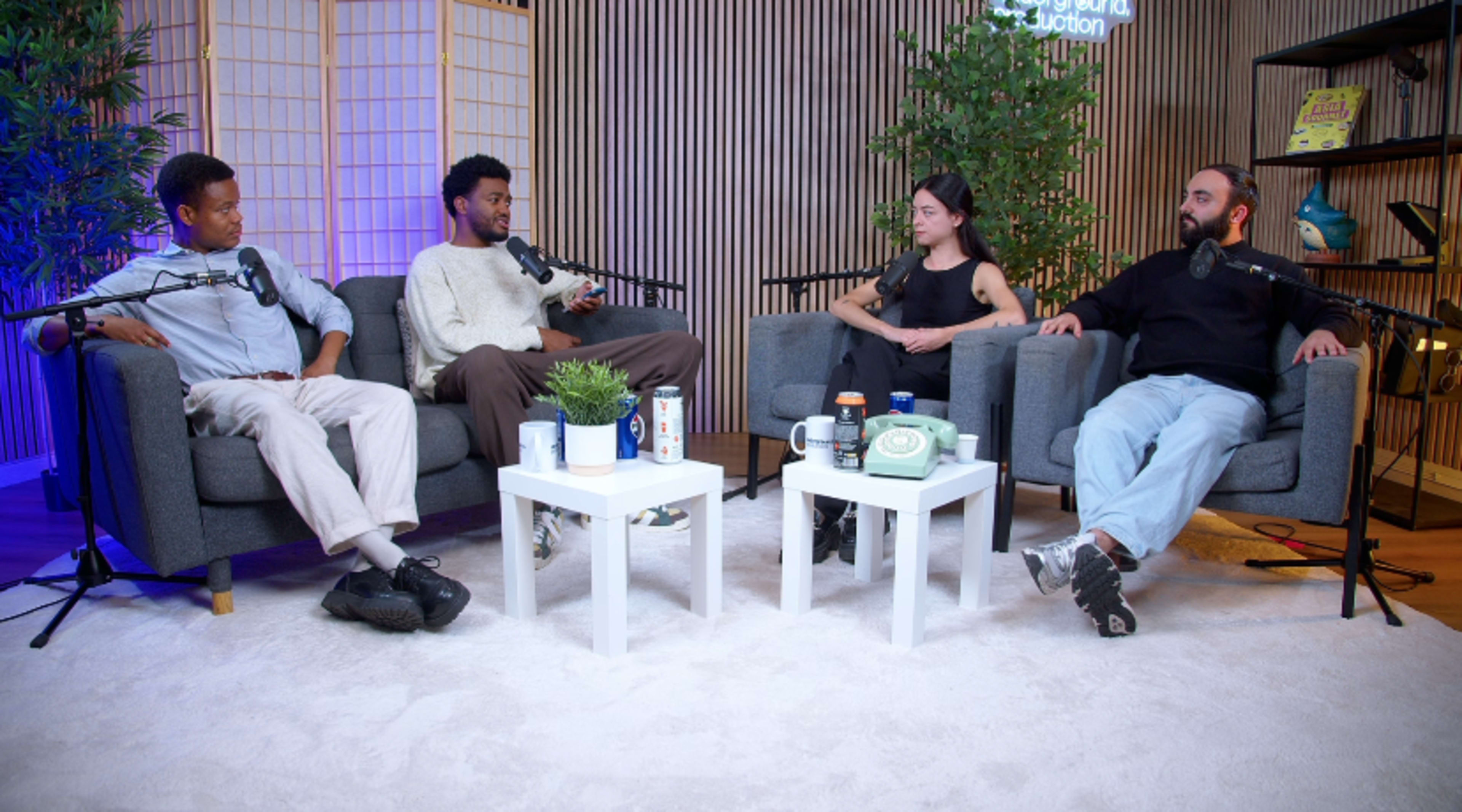 A group of four individuals engages in a discussion while seated in a cozy studio setting with microphones and plants.