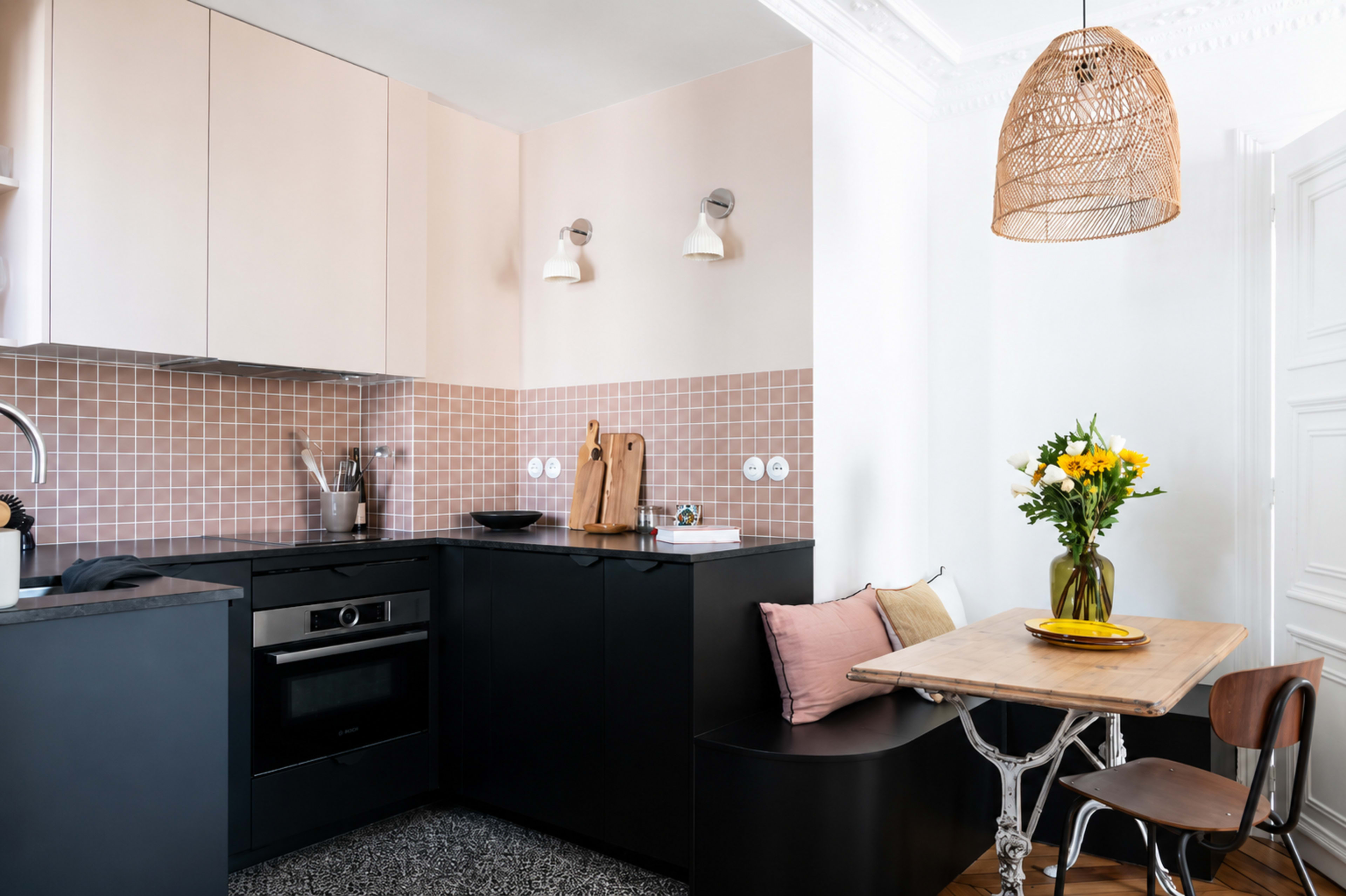The image features a modern kitchen with a black countertop and cabinetry, pale pink wall accents, and a wooden dining table set with a vase of flowers.